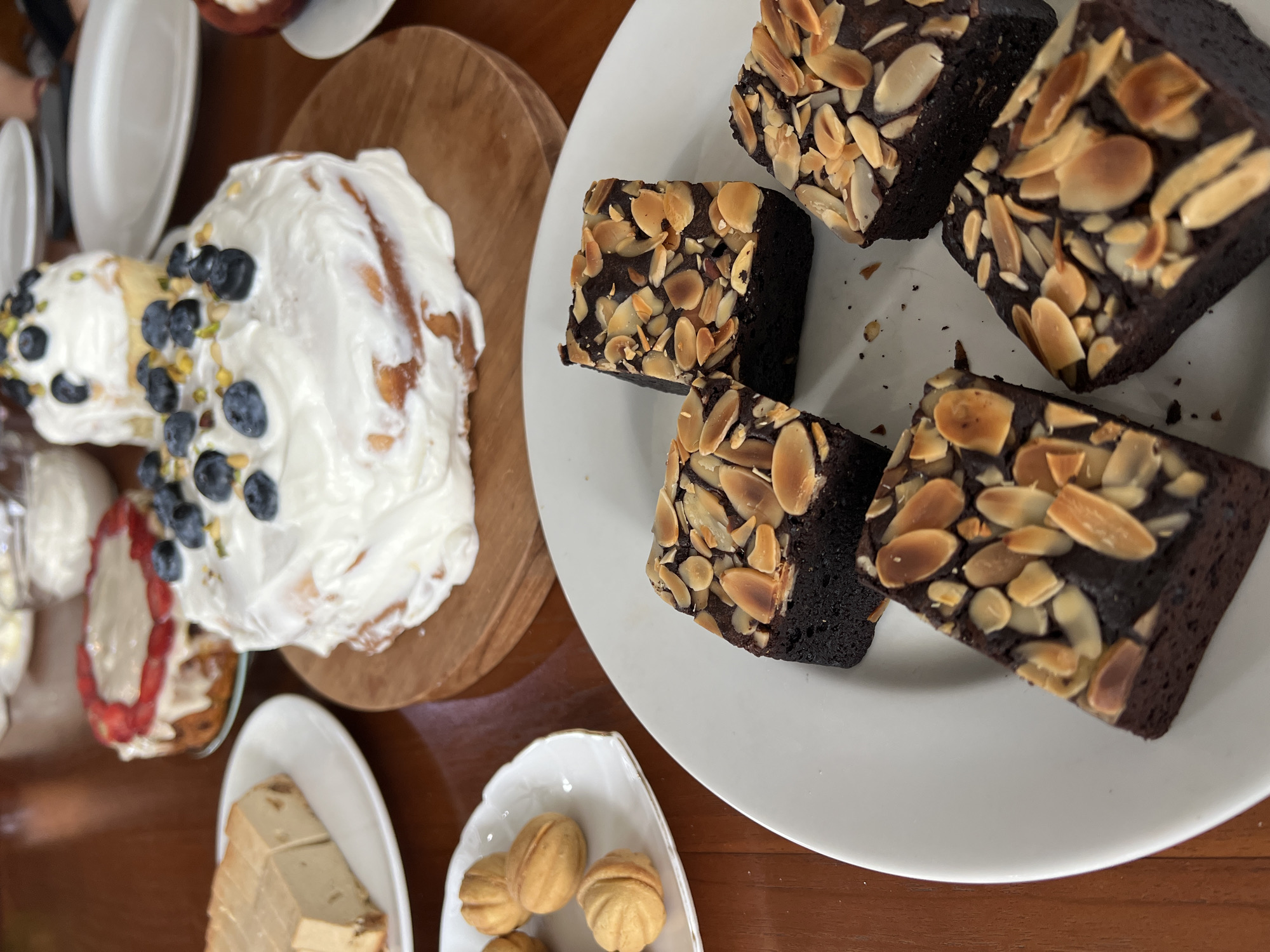 The image shows a top-down view of a table in Singakerta, Indonesia, laden with various desserts. On the left side of the image is a wooden board with a cinnamon roll cake covered in white frosting and decorated with blueberries and chopped pistachios. To the left of the cake are stacks of small, rectangular cakes. Below these are small, nut-shaped desserts arranged on a white, leaf-shaped plate. On the right side of the image, a white plate holds several square brownies topped with sliced almonds. The table has a brown, wooden surface. Several white plates are visible in the upper left corner, suggesting a gathering or meal setting. The focus is on the food, suggesting a culinary theme or a focus on the desserts.