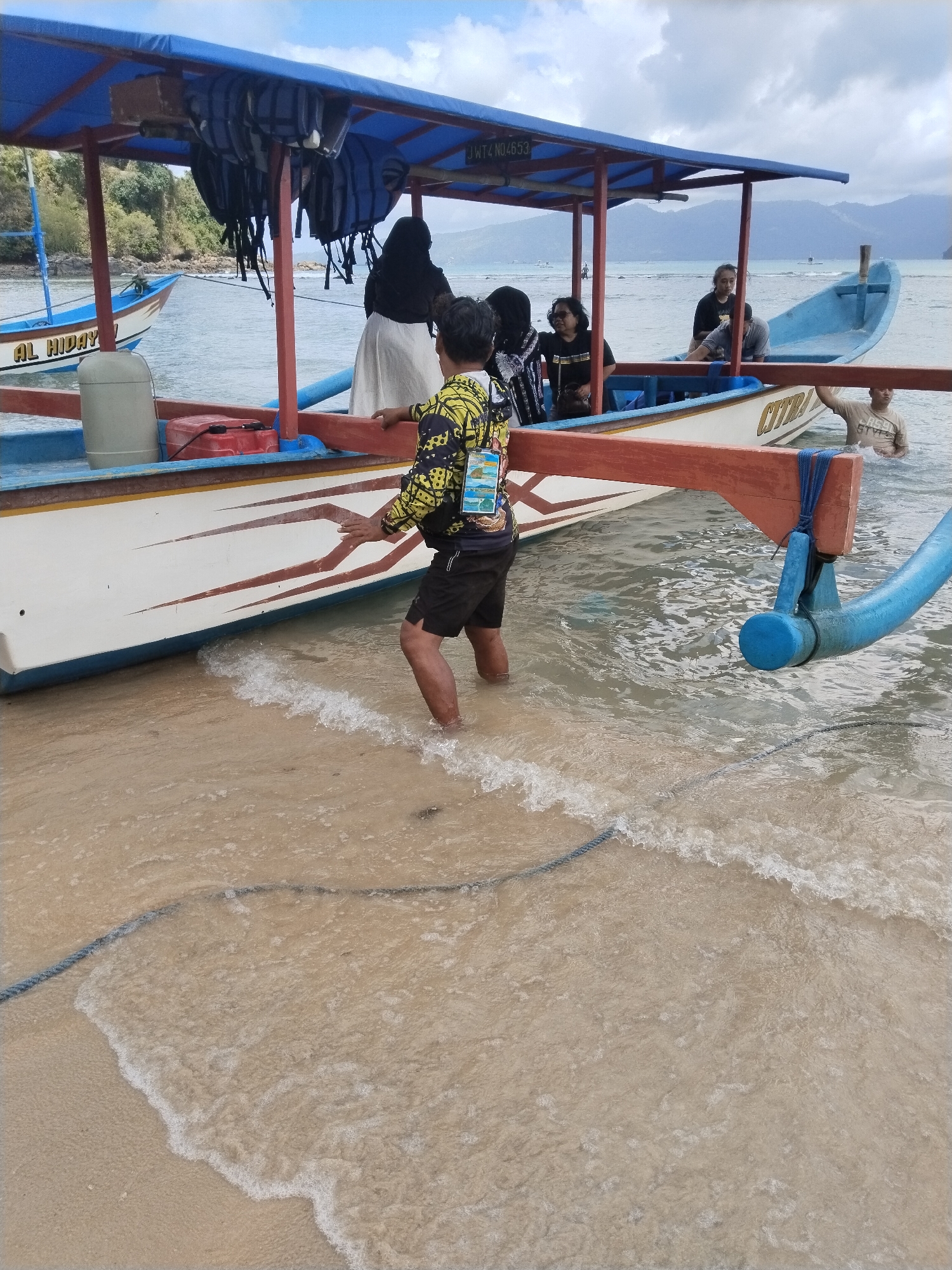 The image captures a scene at the beach in Prigi, Indonesia, where a group of people are interacting with two boats. In the foreground, a person is standing in shallow water, facing away from the camera and towards a boat docked at the shore. The boat has a white hull with red and brown decorative stripes. Life vests are visible hanging from the blue canopy of this boat. Another boat, also blue and white with "AL. HIDAY" written on its hull, is moored slightly further out in the water. Several other individuals are seated or standing within or near the docked boat, some wearing headscarves. The water is clear and shows gentle waves lapping at the sandy shore. In the background, lush green hills and a cloudy sky form the backdrop to the maritime activity.