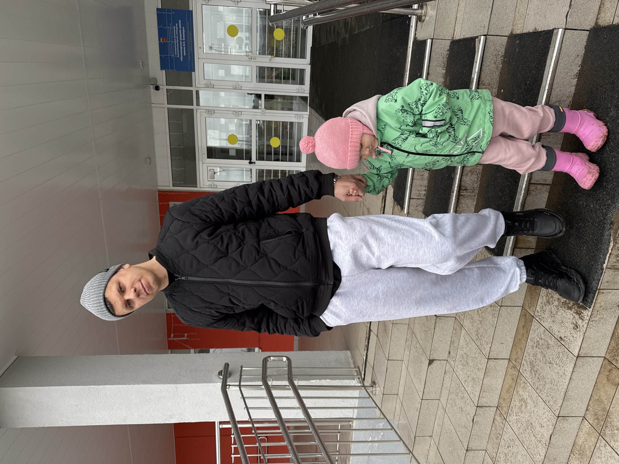 An outdoor scene features a man and a young child holding hands, standing on stone steps in front of a building entrance. The man, positioned on the left, looks towards the camera with a neutral expression. He is dressed in a black quilted jacket, light grey sweatpants, a grey knit beanie, and black sneakers. To his right, the child, who is considerably shorter, is dressed in a light green jacket adorned with a black tiger pattern, light pink pants, a pink knitted bobble hat, and pink rubber boots that show signs of dirt or mud. The child is looking downwards.
The setting appears to be a building entrance with large glass doors and windows. A silver metal handrail extends alongside the steps. The ground is paved with light-colored tiles, and the steps have contrasting dark, textured strips. Visible dampness on the ground and the child's boots suggests wet weather conditions, possibly after rain. The lighting is soft and diffused, indicating an overcast day.
A blue rectangular sign is mounted on the building's facade above the doors. The Russian text on the sign reads: "Муниципальное общеобразовательное учреждение «Константиновская средняя школа имени В.К. Зюканова»". This translates to "Municipal General Education Institution 'Konstantinovskaya Secondary School named after V.K. Zyukanov'", indicating the location is likely a school in the Горный, Russia area.