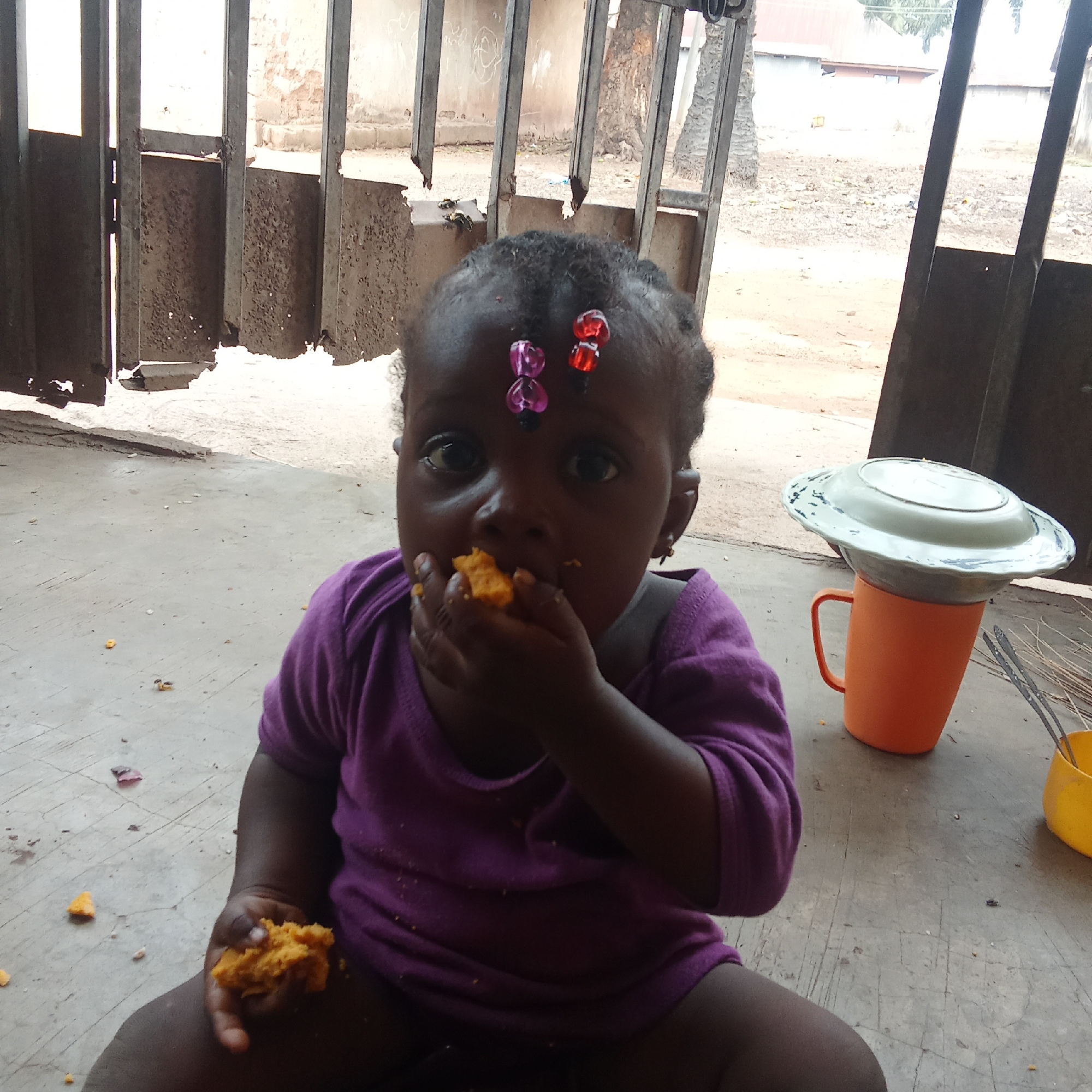 The image depicts a young child, likely between one and two years old, seated on a concrete surface. The child is dark-skinned, wearing a purple shirt, and has decorative beads in their hair. They are holding and eating pieces of an orange-colored food item.
Behind the child, there's a partially visible, weathered metal gate or fence. Adjacent to the child on the right side of the frame is an orange plastic mug topped with a white plate and a yellow container nearby, possibly holding utensils or another food item.
The background visible through the gate suggests a rural or semi-rural setting, with glimpses of buildings and vegetation.
Based on the prompt, the location is presumed to be in Akwanga, Nigeria. The setting appears to be an outdoor, possibly residential or semi-public space.