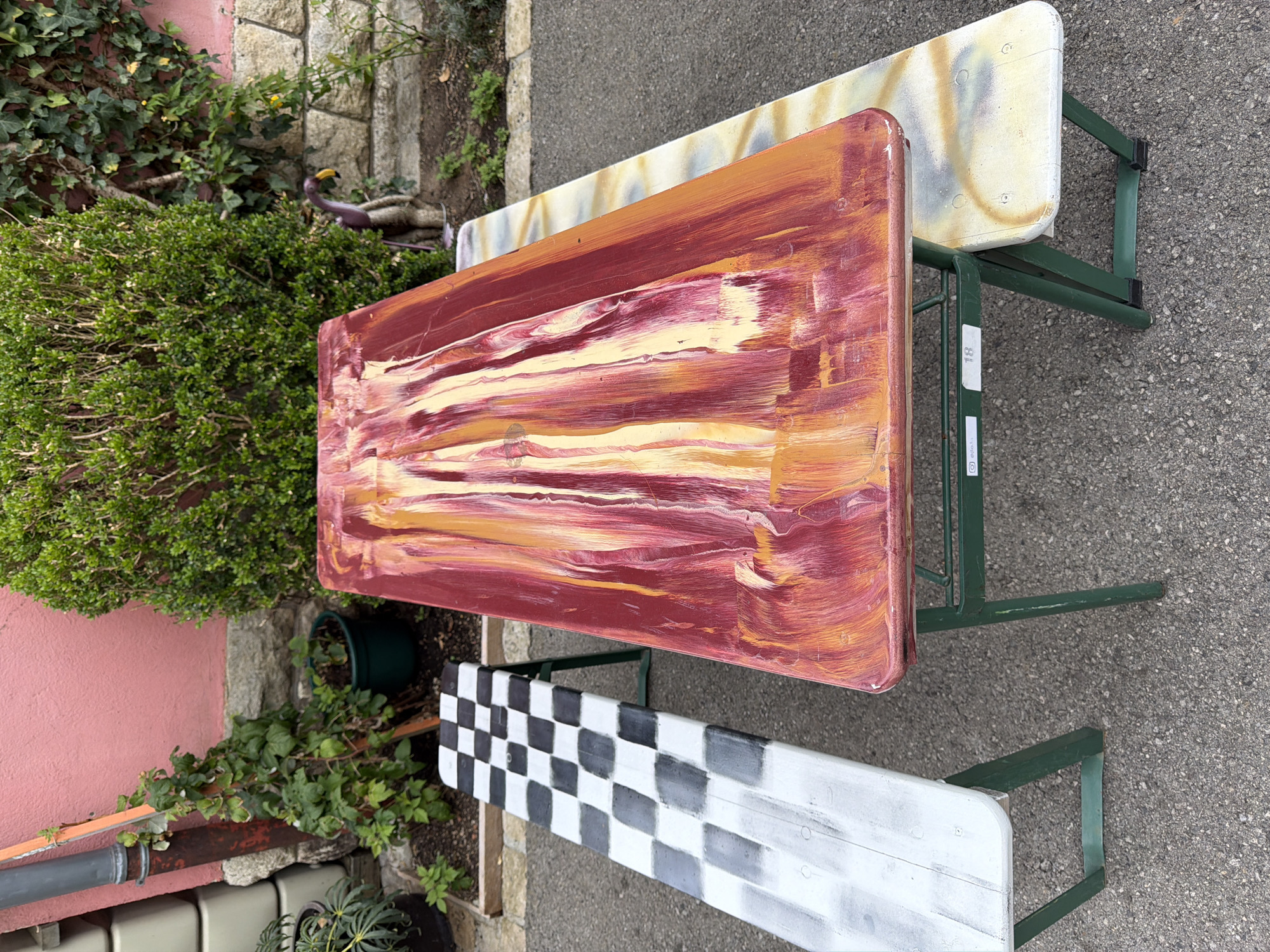 The image depicts an outdoor scene in Esch-sur-Alzette, Luxembourg, focusing on two beer garden benches and a table. The central object is a rectangular table with a top painted in a chaotic, abstract pattern of red, orange, and yellow streaks, resembling dripping paint or a flame effect. It rests on dark green metal legs. To the left of the table is a bench painted with a black and white checkered pattern, transitioning to a white and gray gradient. This bench also has dark green metal legs. To the right of the table, another bench is visible, its top painted with a light yellow and gray abstract pattern. The scene is set outdoors on an asphalt surface. In the background, a pink wall, a dense green bush, and a stone wall can be seen. A drainpipe runs down the pink wall, and some greenery is present around the base of the walls. A small, purple and brown toy flamingo is placed near the bush. The overall impression is one of a decorated outdoor seating area, possibly in a garden or yard. There are no people or animals actively engaged in any action within the frame.