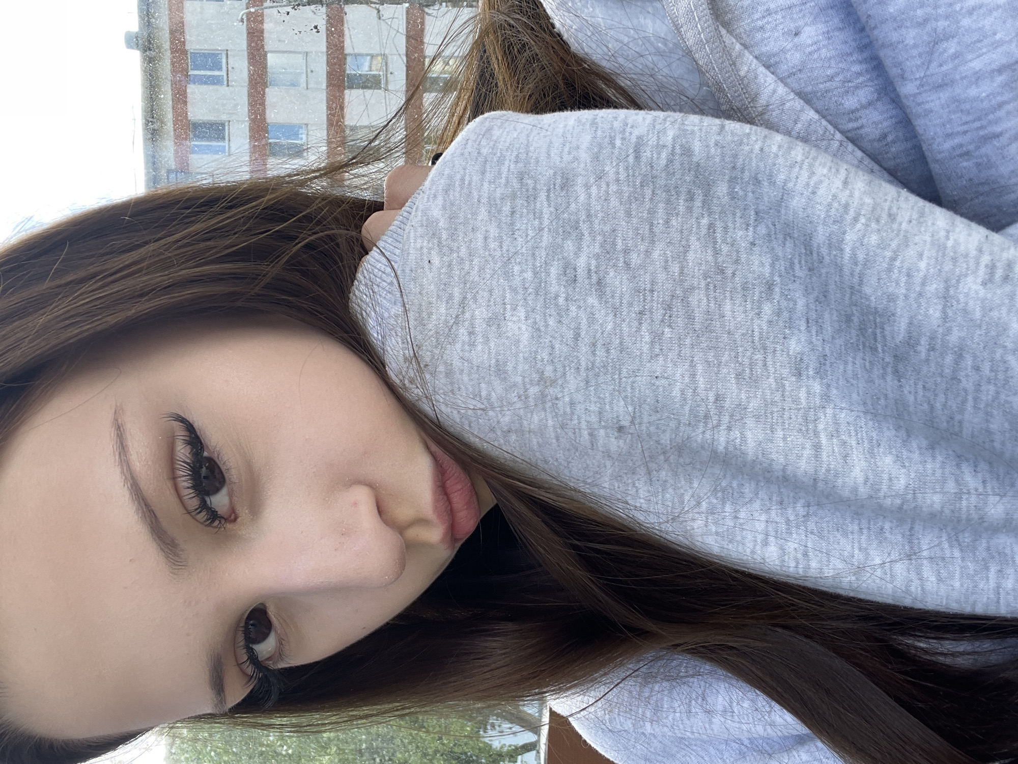 A close-up, slightly rotated portrait shows a young woman with brown hair and brown eyes. She is wearing eyelash extensions. She is leaning against a gray, textured sweatshirt. A building exterior can be seen blurred in the background. Based on visual assessment, the building appears to be a multi-story residential structure with a repetitive window pattern. The location is in the city of Chaikovsky, Russia. The environment suggests an overcast day.