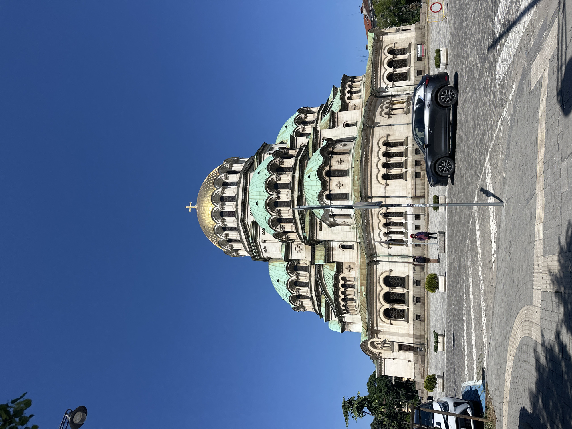 The image depicts the Alexander Nevsky Cathedral in Sofia, Bulgaria, on a sunny day. The cathedral's architecture, including its domes and façade, is prominently featured. A dark-colored car is parked on the pavement beside the cathedral. Pedestrians can be seen on the walkway near the building. The ground is paved with stone or concrete. The sky is a clear blue.