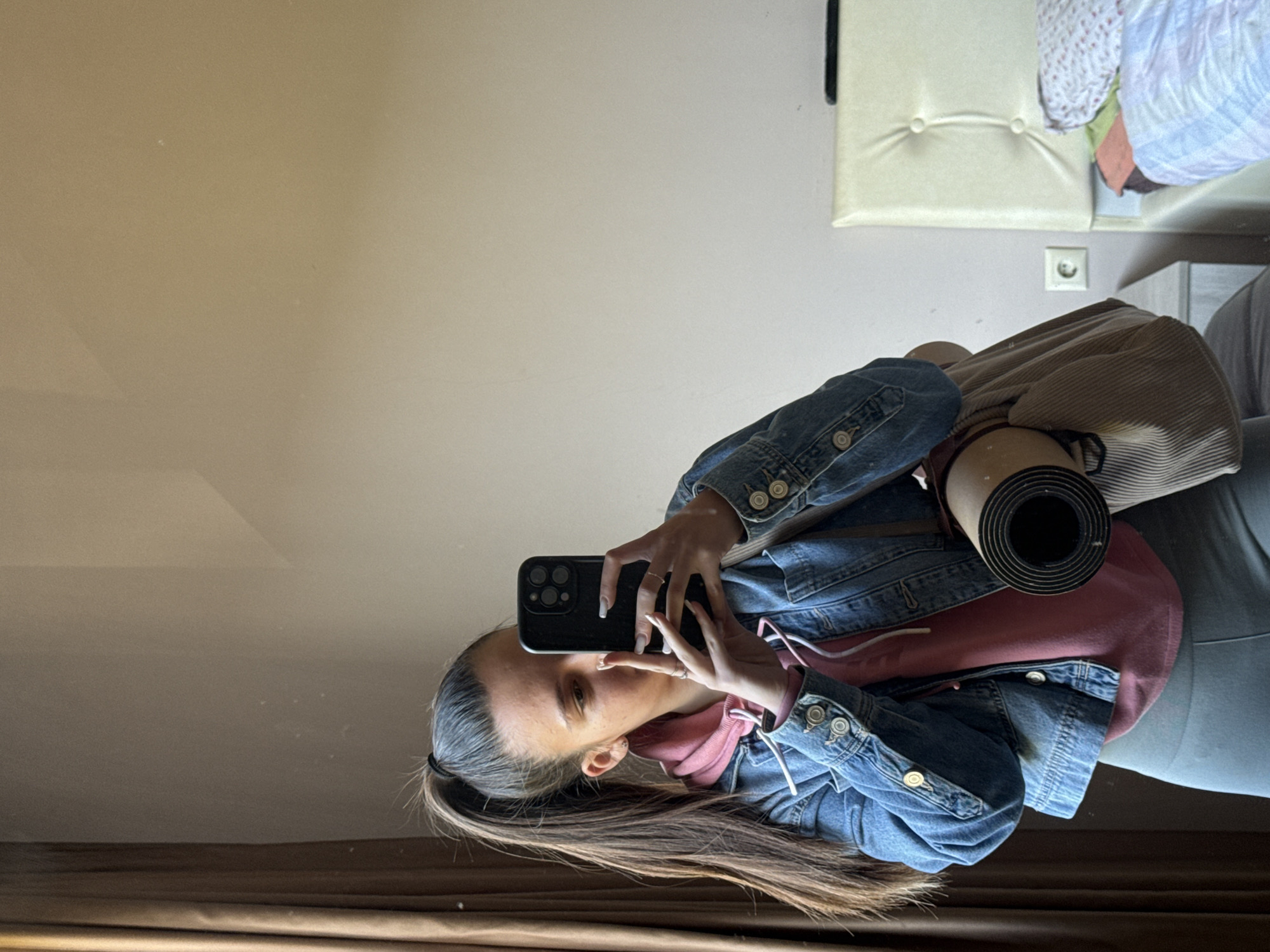 A woman in a denim jacket and pink hoodie is taking a selfie in a mirror. She is holding a black smartphone in her hands. A yoga mat is visible near her shoulder. The background includes a cream-colored wall, a padded bench, and a European-style electrical outlet. The setting appears to be indoors, possibly in a home. The exact location within Varna, Bulgaria cannot be determined.