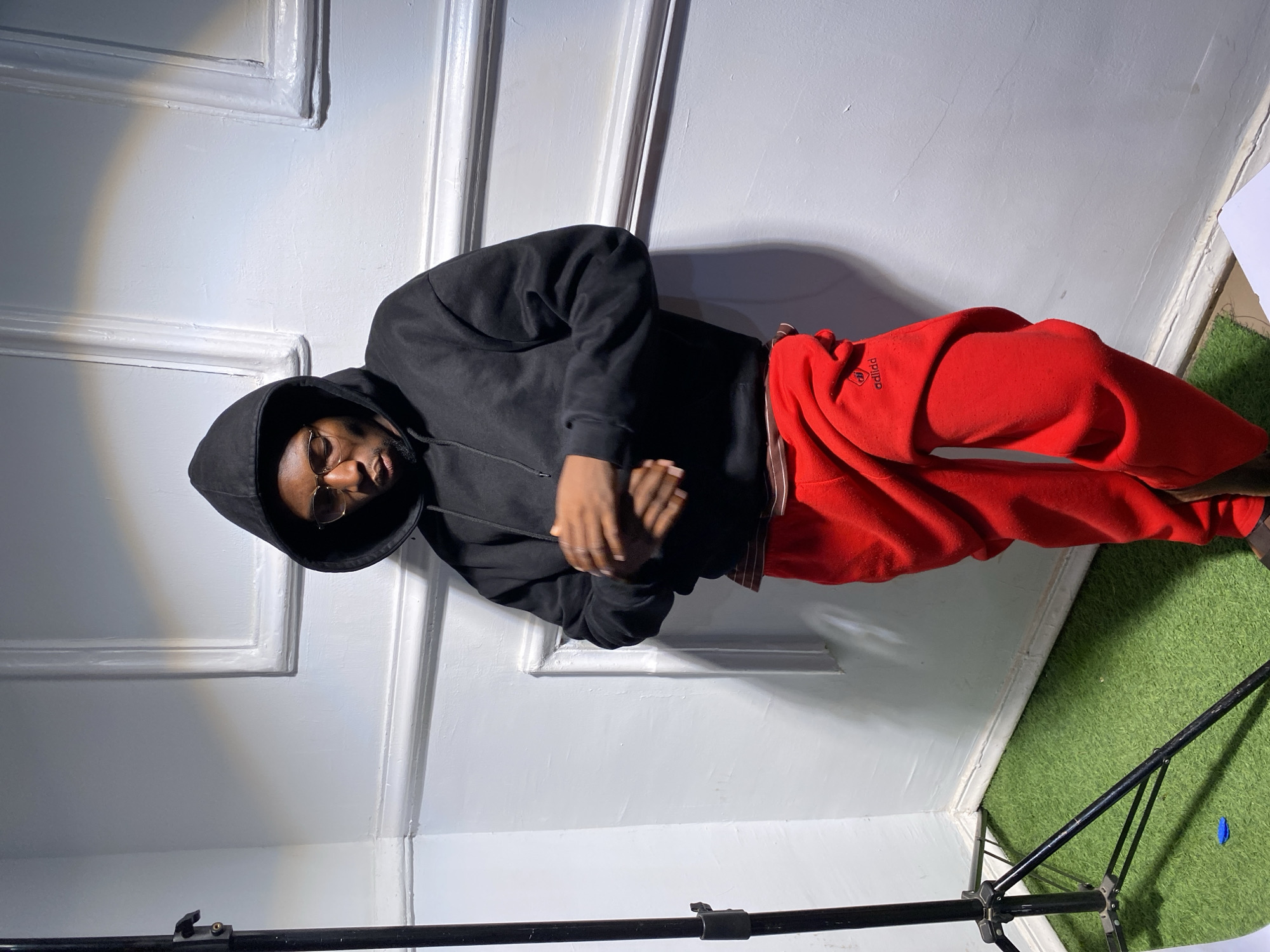 The image shows a young Black man posing for a photograph indoors in Barakin Akawo, Nigeria. He is wearing a black hooded sweatshirt with the hood up and bright red sweatpants. The sweatpants have white text on the left thigh, which appears to be a stylized or slightly misspelled "adidda" logo (likely referencing Adidas). The man wears eyeglasses with thin frames and has a neutral expression, looking slightly downwards with his hands clasped in front of him.
He is standing against a white wall featuring decorative architectural molding, creating a panelled backdrop. The floor beneath him is covered with green artificial turf. A strong diagonal shadow is cast on the white wall behind the man's head, suggesting a direct light source from the side. In the bottom right foreground, a section of a black tripod or lighting stand is visible, indicating a studio-like setup. A small, crumpled blue object rests on the green turf near the stand. The overall scene suggests a casual photoshoot in a controlled environment.
