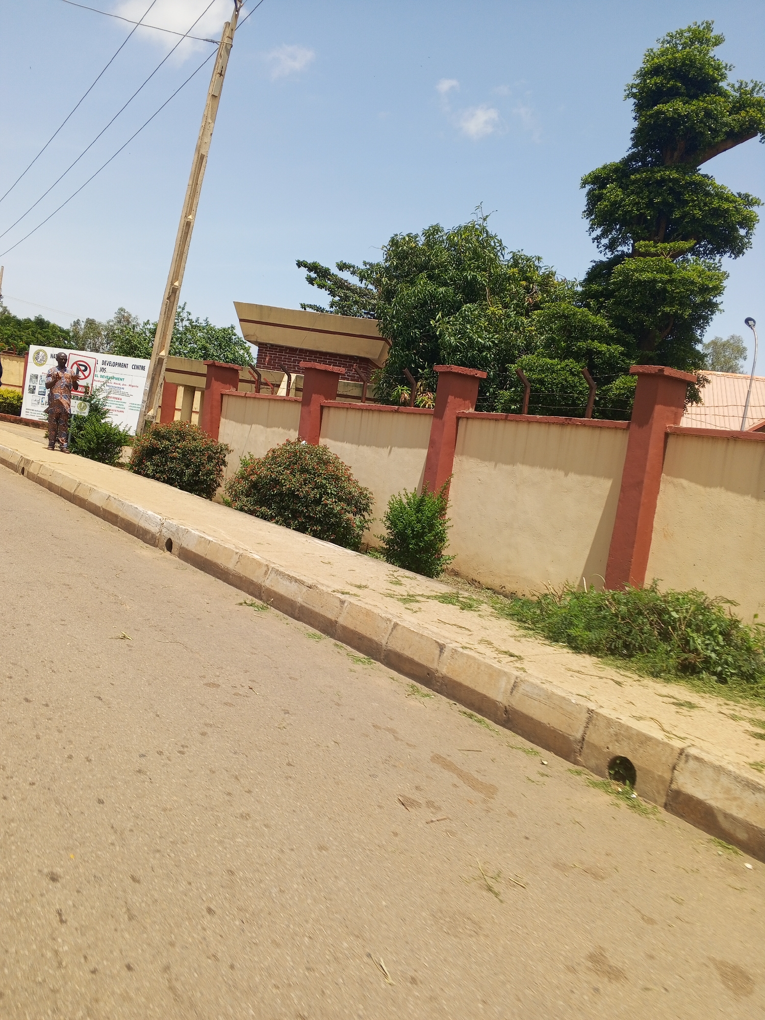 The image shows a street scene in the city of Jos, Nigeria, near a national development center. A paved road with a concrete curb and drainage gutter is in the foreground, with some grass clippings visible along the edge of the road and sidewalk. A sidewalk runs alongside a beige-colored wall topped with red brick pillars. Behind the wall, lush green trees and bushes are abundant. On the sidewalk, a man in a colorful patterned outfit stands next to a sign that reads "NATIONAL DEVELOPMENT CENTRE JOS". The sky is blue with scattered clouds, and a utility pole with power lines extends upwards. The overall impression is an urban environment with a strong presence of greenery.