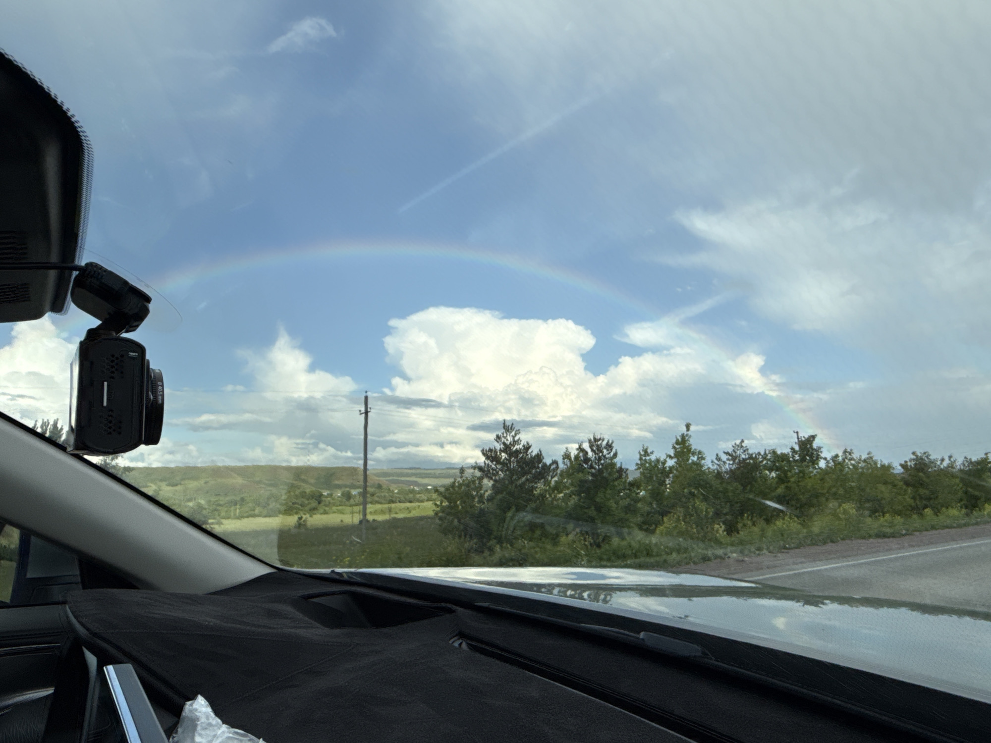 The image depicts a rural landscape viewed through the windshield of a vehicle in Субханкулово, Russia. The sky, occupying the upper two-thirds of the frame, is blue with scattered white cumulus clouds. A prominent rainbow arcs across the sky from the left to the right side, above the horizon. A faint linear cloud or contrail is also visible in the upper central sky.
In the immediate foreground, the interior of the vehicle is visible. The underside of a black rearview mirror assembly is partially seen at the top-left, and a black dashcam with a cylindrical lens (labeled "Ø40.5 mm") is mounted to the windshield below it. The dashboard, covered with a dark, textured fabric mat, occupies the lower portion of the frame. Reflections of the sky and clouds are present on the windshield, particularly at its base.
The exterior landscape features rolling green hills in the distance, covered with sparse vegetation. A cluster of dark green trees is visible in the midground, bordering the right side of a paved road with a light-colored shoulder. A tall utility pole with a crossbar stands upright on the left side of the midground. The ground appears damp, with reflections of the sky visible on a wet surface near the road in the foreground. No people are visible in the scene.