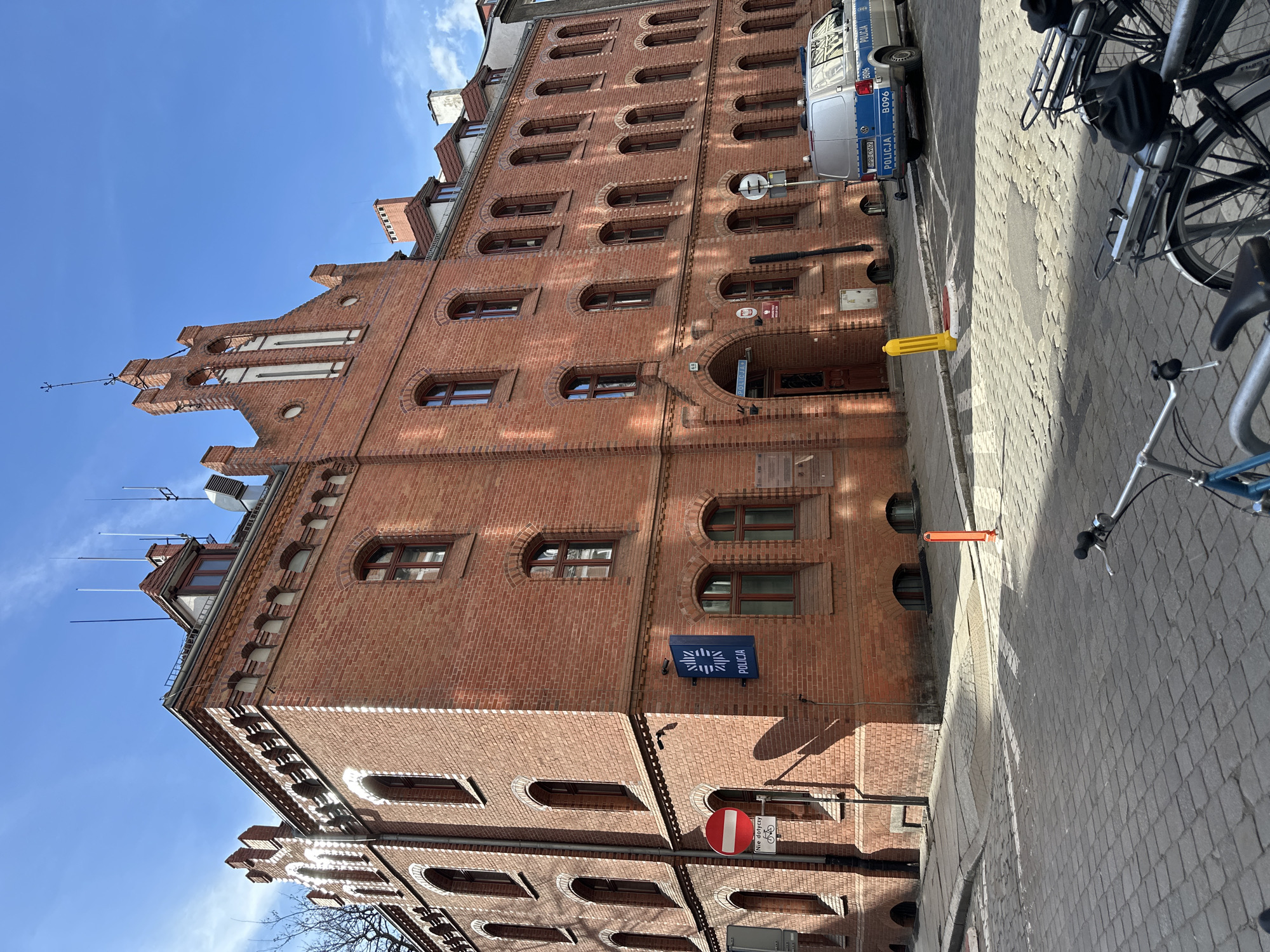 The image shows a brick building under a clear blue sky, identified as the Police Department in Wrocław, Poland, due to the "POLICJA" (Police) sign on its facade. The building features arched windows and decorative brickwork. A police van is parked on the street beside the building. Bicycles are also visible on the sidewalk. A "no entry" traffic sign is attached to the building. Several yellow and orange traffic barriers are present near the sidewalk. The pavement is paved with interlocking stones.