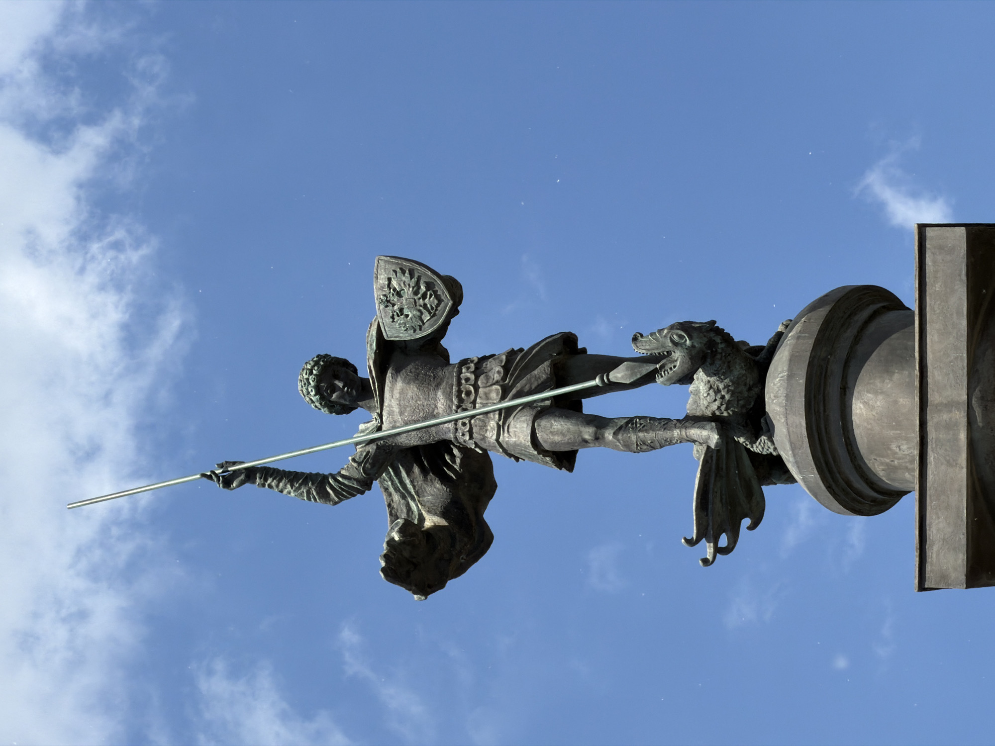 A bronze statue, likely depicting Saint George slaying the Dragon, stands prominently against a bright blue sky with scattered white clouds. The monumental sculpture features a knightly figure, clad in intricate armor and a helmet, captured in a dynamic pose. He holds a long, slender spear in his hands, thrusting it forward and slightly downwards. On his left arm, he bears a shield embossed with a detailed emblem, possibly a coat of arms.
Below the figure's right leg, a fierce-looking dragon with textured scales and visible wings is depicted, its head turned back. The entire sculptural group, including the knight and the dragon, rests upon a robust, multi-tiered bronze pedestal with both rounded and angular sections. The statue's dark, patinated bronze contrasts sharply with the vivid blue of the sky, indicating a clear, sunny daytime. The low-angle perspective emphasizes the heroic scale and dramatic action of the scene. The intricate details of the armor, the dragon's features, and the shield's emblem are clearly visible. This public monument is located in Moscow, Russia. No text is visible on the statue or pedestal.