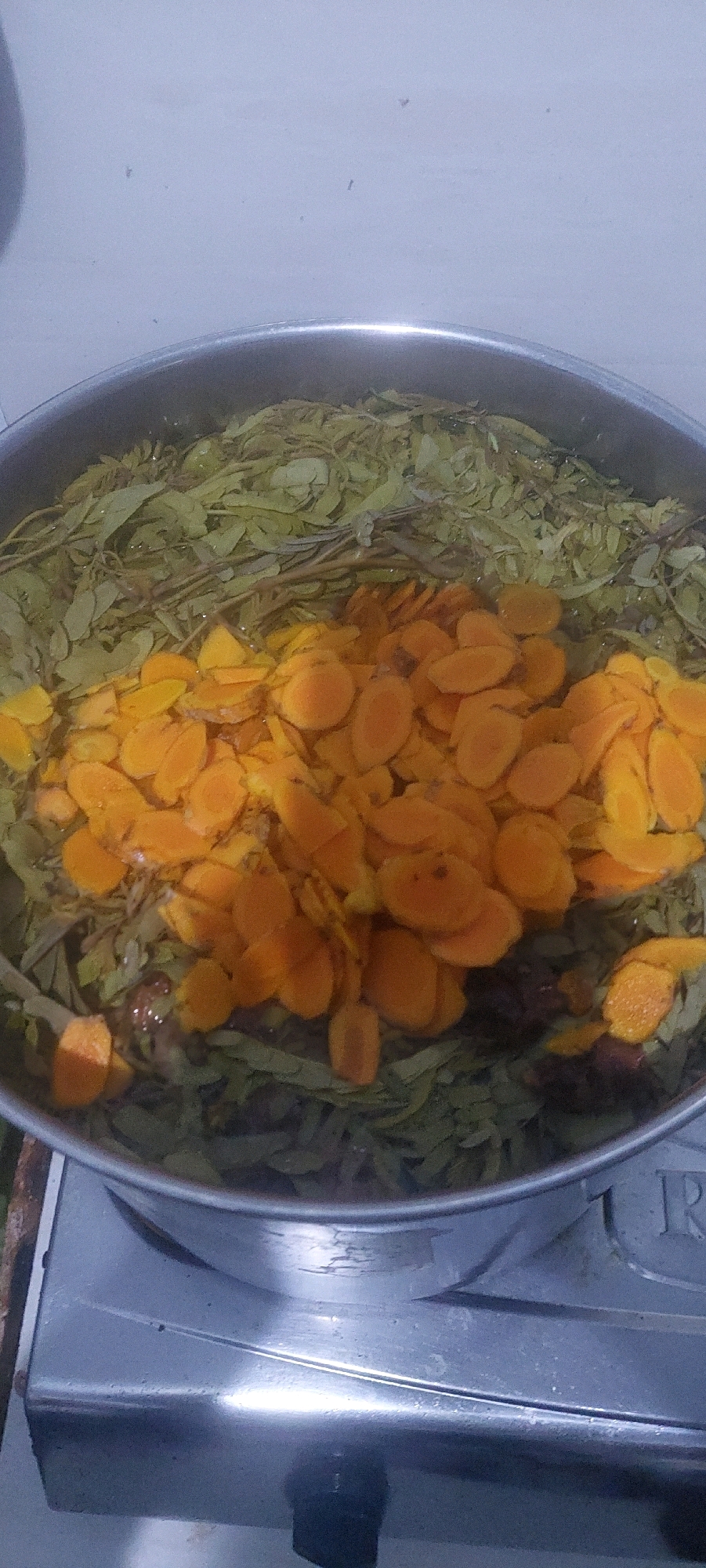 In Lumajang, Indonesia, a close-up shot focuses on a pot of simmering herbs and sliced turmeric on a stovetop. The pot is filled with green leaves and branches, likely herbs, submerged in water. A thick layer of bright orange, thinly sliced turmeric is piled on top of the herbs, partially obscuring them. The pot is made of stainless steel and appears to be a standard kitchen pot. The stovetop is a metallic, possibly stainless steel, surface with a burner visible at the bottom of the frame. A partial view of what appears to be a gas stove knob is present at the very bottom. The lighting is dim, casting a slight shadow on the stovetop. The background is a plain, light-colored wall.