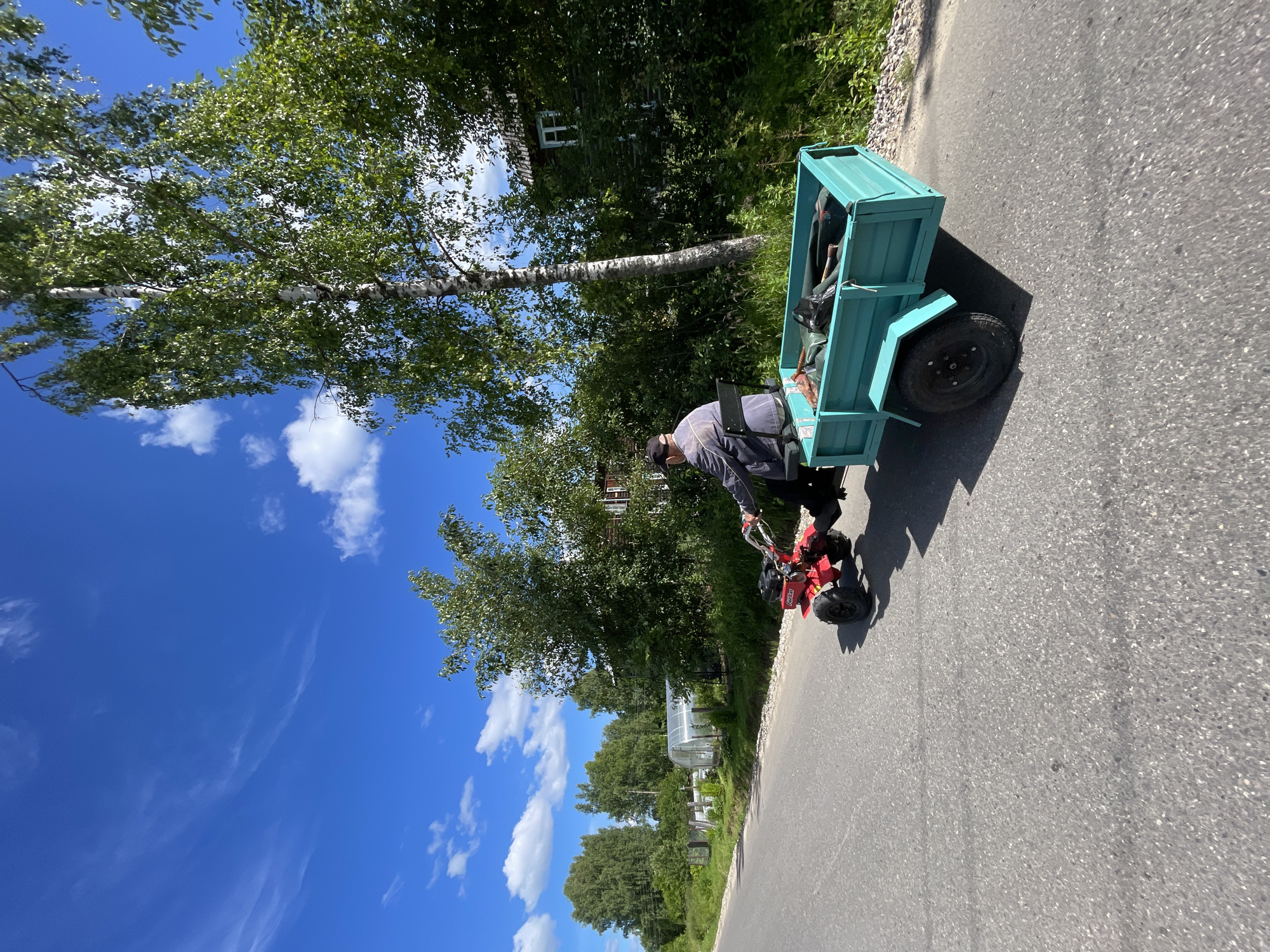 A person is riding a red tiller with a turquoise trailer attached, on an asphalt road. The person is wearing a dark jacket and a baseball cap. The trailer contains gardening tools and a black plastic bag. The scene is set in a rural area with trees and houses on the right side of the road, and greenhouses and fences on the left. The sky is blue with scattered white clouds. The image appears to have been taken in a location like Заречный, Russia, given the rural setting and architectural style of the houses.