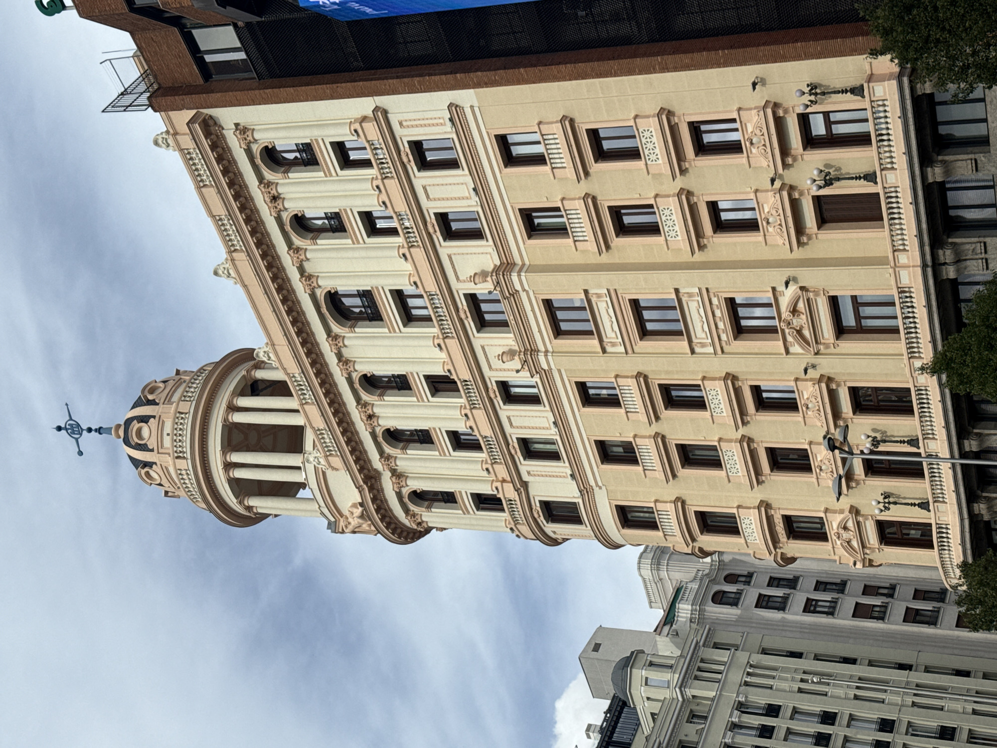 The image shows a view of the Edificio Metrópolis, a historic building in Madrid, Spain. The building features a distinctive cylindrical corner tower topped with a dome and a statue of a winged Victory. Its architecture is characterized by Beaux-Arts style with ornate detailing, including decorative carvings, columns, and window treatments. The building is cream-colored with brown window frames and accents. The sky is cloudy. Other buildings are visible in the background, providing context for its urban location.