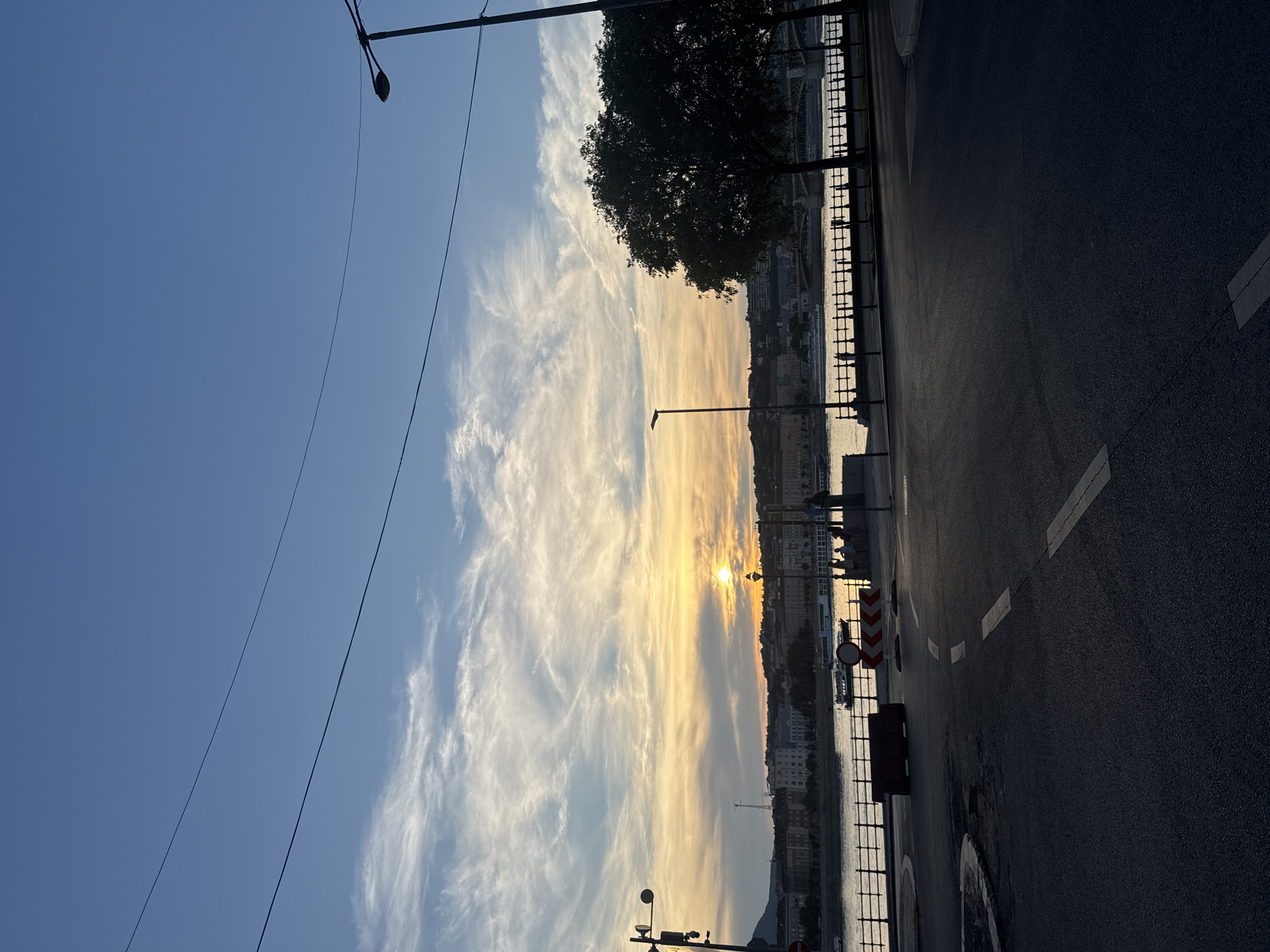 The image shows a sunset over the Danube River in Budapest, Hungary. The sky is filled with wispy clouds, tinged with orange and yellow from the setting sun. The sun is low on the horizon, casting a warm glow across the water. In the foreground, there is a paved road with white lane markings. A black lamppost stands on the side of the road, and there are some traffic cones and barriers. Across the river, the cityscape of Budapest is visible, with buildings and a bridge. The overall mood is peaceful and serene, with a sense of tranquility. There is no visible text in the image.