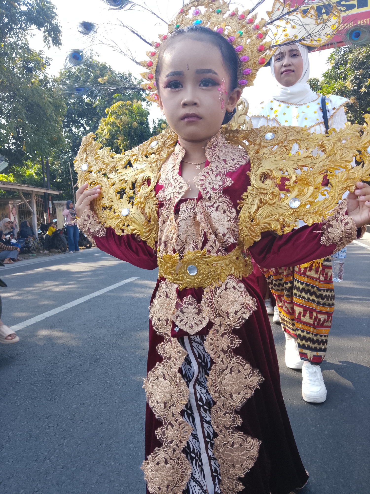 In Buduan, Indonesia, a young girl in elaborate traditional attire, adorned with peacock feathers and gold detailing, participates in a parade. She wears a deep red velvet dress with gold lace accents and a striking golden headdress. Beside her, a woman in a white hijab and a patterned outfit also participates. The parade takes place on an asphalt road, with trees and buildings visible in the background. Spectators are gathered along the side of the road, some sitting and others standing near motorcycles. The sunlight casts shadows, indicating a bright, sunny day.