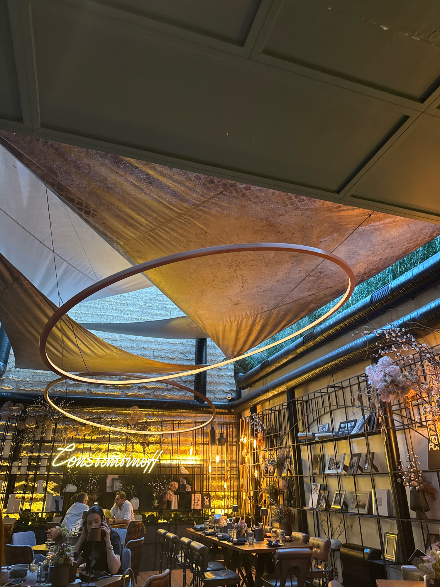 The image shows the interior of a restaurant with a modern, somewhat industrial design. The ceiling features an abstract canopy of fabric and a large, circular pendant light with integrated LED lighting. The walls are adorned with shelves displaying framed photographs, books, and decorative floral arrangements, creating a gallery-like atmosphere.
In the foreground, several tables are set for dining, with chairs and place settings visible. A woman in a black shirt is seated at one of the tables, holding a phone to her ear. Other patrons are seated at tables in the background, some engaged in conversation. A neon sign spelling out "Constantinoff" in script is prominently displayed on the wall. The overall lighting is warm and ambient, with exposed light bulbs and the glow from the neon sign contributing to the mood. The restaurant appears to be located in Sofia, Bulgaria, as suggested by the "Constantinoff" neon sign.