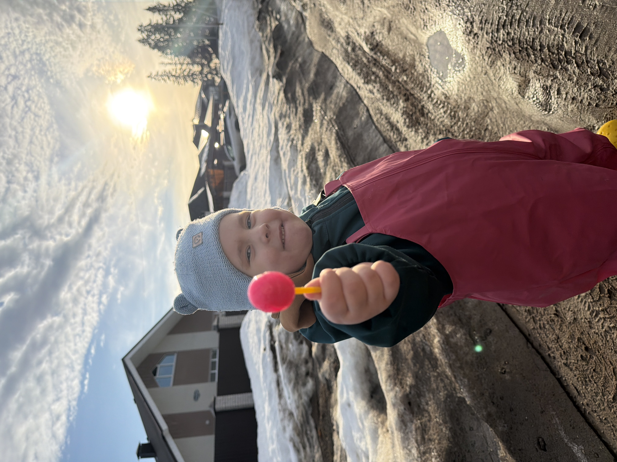 The image depicts a young child, approximately 3-5 years old, standing outdoors in Chaikovsky, Russia. The child is positioned centrally, facing forward, and looking directly at the camera with a slight smile. The child wears a light blue knitted hat with two small ear-like protrusions, a dark green jacket, and bright pink waterproof overalls. The child's right hand is extended towards the viewer, holding a prominent bright pink, spherical lollipop on a yellow stick.
The immediate ground consists of irregular patches of dirty melting snow, exposed muddy earth, and tire tracks. Small puddles are visible, reflecting sunlight. In the background to the left, a two-story residential house with light beige and brown exterior walls, a dark peaked roof, and a chimney is visible. A dark fence with brown brick pillars stands in front of this house. To the right, the sun is low in the sky, emitting a strong golden light and lens flare. Several tall evergreen trees are silhouetted against the bright sky, with additional residential buildings visible further in the distance, partially obscured by snowdrifts. The sky is blue with scattered white, wispy clouds. The scene suggests a transition season, likely late winter or early spring, given the presence of melting snow and low sun angle.