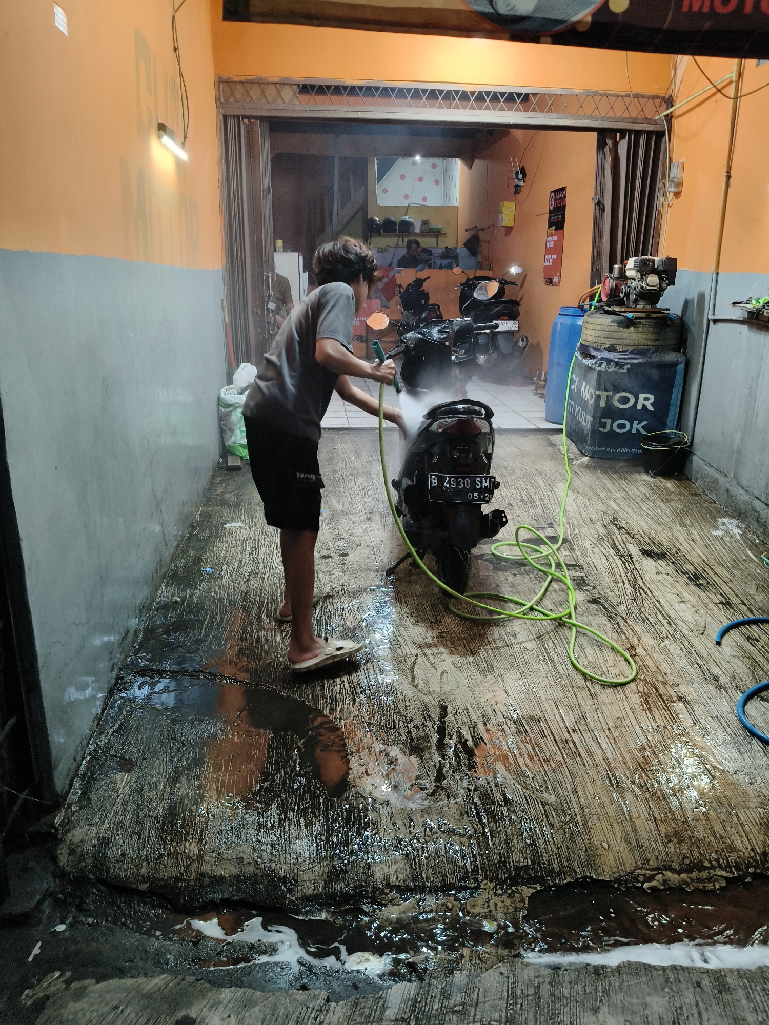 In South Tangerang, Indonesia, a young person is washing a black motorcycle in a covered outdoor area. The person, wearing a grey t-shirt and black shorts, is using a green hose to spray water onto the rear of the motorcycle, which has a license plate reading "B 4930 SMT 05-24". The ground is wet and appears to be concrete with some debris and puddles of water, suggesting a car wash or service area. In the background, other motorcycles are parked, and various equipment, including a blue water tank and a compressor, are visible. The walls are painted in orange and grey, and a fluorescent light fixture is mounted on the ceiling. The overall scene depicts a routine vehicle maintenance activity in an urban Indonesian setting.
