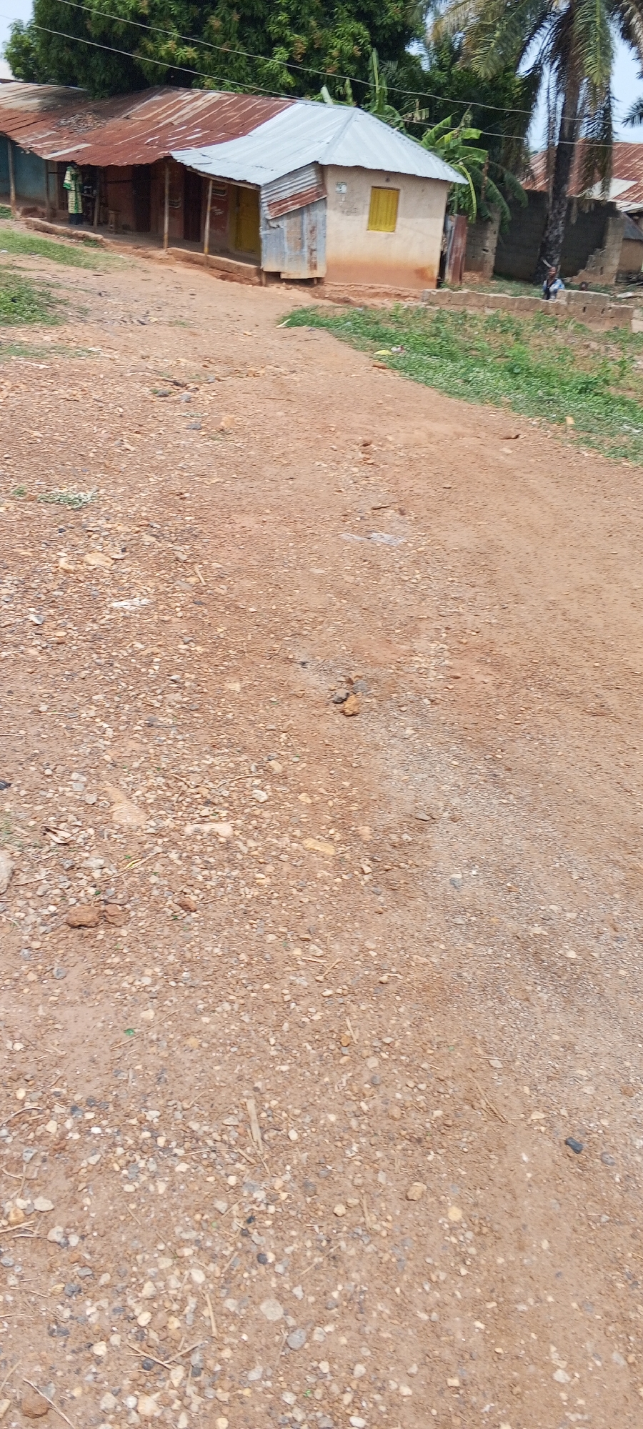 The image shows a dirt road in the Unknown location of the city of Unknown. The road is covered in small rocks and pebbles. In the background, there are several houses with corrugated metal roofs. Some trees and vegetation are visible in the background as well. There are no people visible in the foreground. One person is seen in the distance by a house.