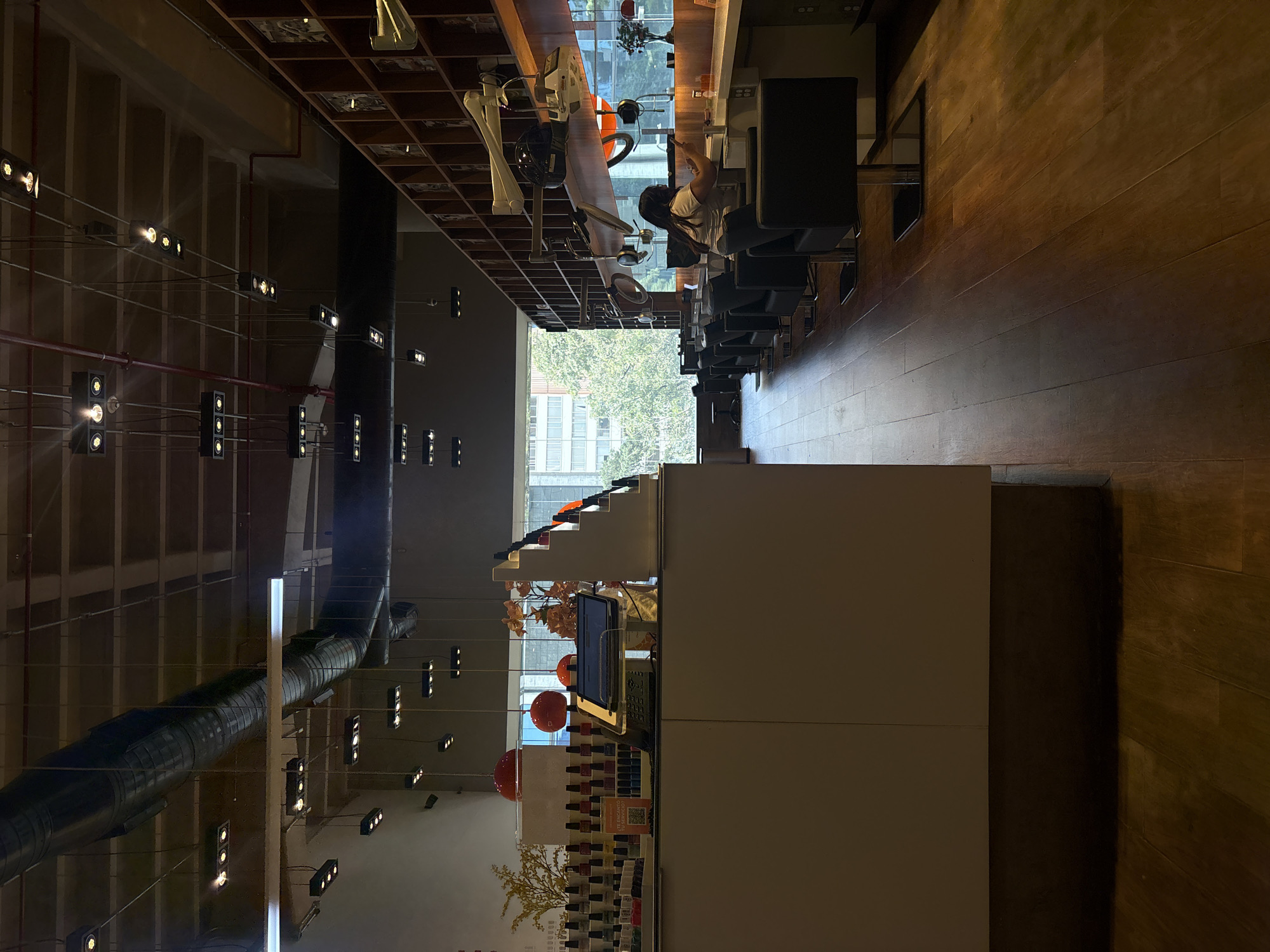 The image shows the interior of a modern nail salon or beauty parlor in Mexico City. The scene is captured from a slightly elevated perspective, looking down the length of the salon. The flooring is made of polished dark wood planks, and the ceiling features exposed industrial ductwork and a grid of recessed track lighting.
In the foreground, to the left, is a white counter with a tablet device and a cash register, suggesting a reception or checkout area. Shelves behind the counter are stocked with rows of nail polish bottles in various colors. Decorative elements include two large, round red pendant lights hanging from the ceiling and some floral arrangements.
The middle ground of the image is dominated by a row of black, modern salon chairs facing a long counter. A young woman with dark hair, wearing a light-colored t-shirt, is seated at one of these stations, facing away from the camera, and appears to be working on her phone or a client's nails.
Along the right side of the salon, a wall is lined with wooden shelves, displaying various products and framed images. Suspended above the counter are several salon-style hairdryers, suggesting this might also be a hair salon. Large windows in the background offer a view of trees and buildings, indicating an urban setting. The overall impression is a clean, stylish, and well-lit commercial space.