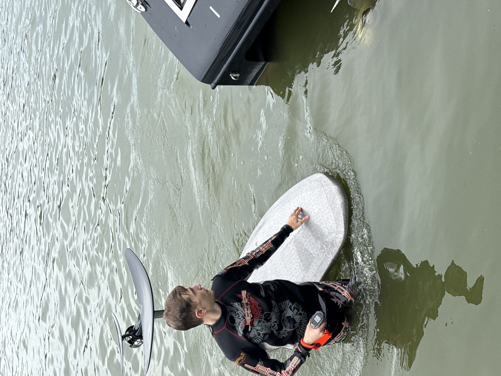 An athletic male in a black and red wetsuit with a "Jetribe" logo stands in murky water, holding a foil board and a black electronic device. He is looking to his right. A dark-colored boat is partially submerged to the right of the frame. The water is rippled, suggesting movement. The scene is consistent with water sports activities in a body of water like the Votkinsk Reservoir, located in the city of Votkinsk, Russia.