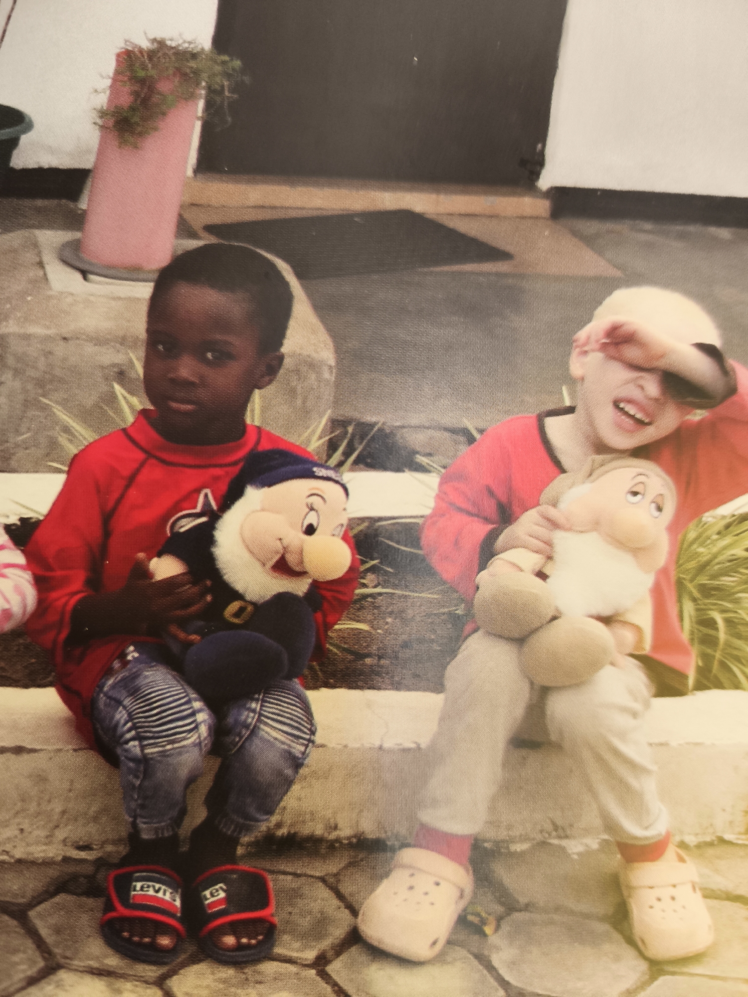 The image depicts two young children sitting on concrete steps outdoors. The child on the left, with darker skin and hair, is wearing a red long-sleeved shirt, patterned blue jeans, black socks, and red and black slide sandals with the "Levi's" logo. This child is holding a plush toy resembling a dwarf with a blue hat and beard. The child on the right, with lighter skin and hair, is wearing a red long-sleeved shirt, light-colored pants, pink socks, and beige clogs. This child is holding a plush toy of a dwarf with a beard and is shielding their eyes with their hand, with their mouth open in a yell. The background includes a pink cylindrical object with plants on top, a dark mat, and a white wall. The image appears to be taken on a sunny day. The location in Passignano sul Trasimeno, Italy, is not discernible from the visual information provided in the image.