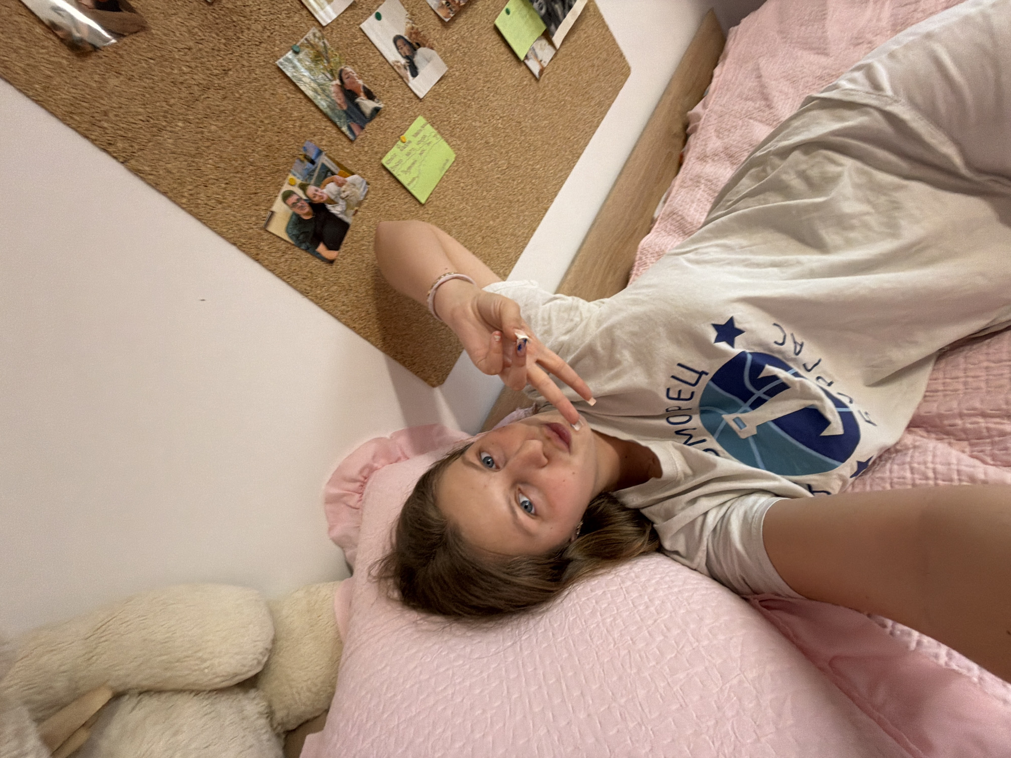A young woman is captured in a selfie, lying on her back on a bed, looking directly at the camera with a relaxed expression. She has light brown hair and blue eyes, and is making a peace sign with her right hand. She wears a light-colored t-shirt featuring a blue and white logo with a star, and Cyrillic text that partially reads "МОДЕЛ" (MODEL) and "СПОРТ" (SPORT) or "СПОРТАК". A light-colored bracelet is visible on her right wrist, and her nails have subtle polish.
The bed is covered with pink, textured bedding, and she rests her head on a matching pink pillow. A large, fluffy, off-white stuffed animal is partially visible to her left. In the background, a cork board is mounted on a light-colored wall, displaying several pinned photographs of various individuals and a small green sticky note with illegible handwritten text. The scene depicts a casual indoor setting, likely a bedroom in Burgas, Bulgaria.