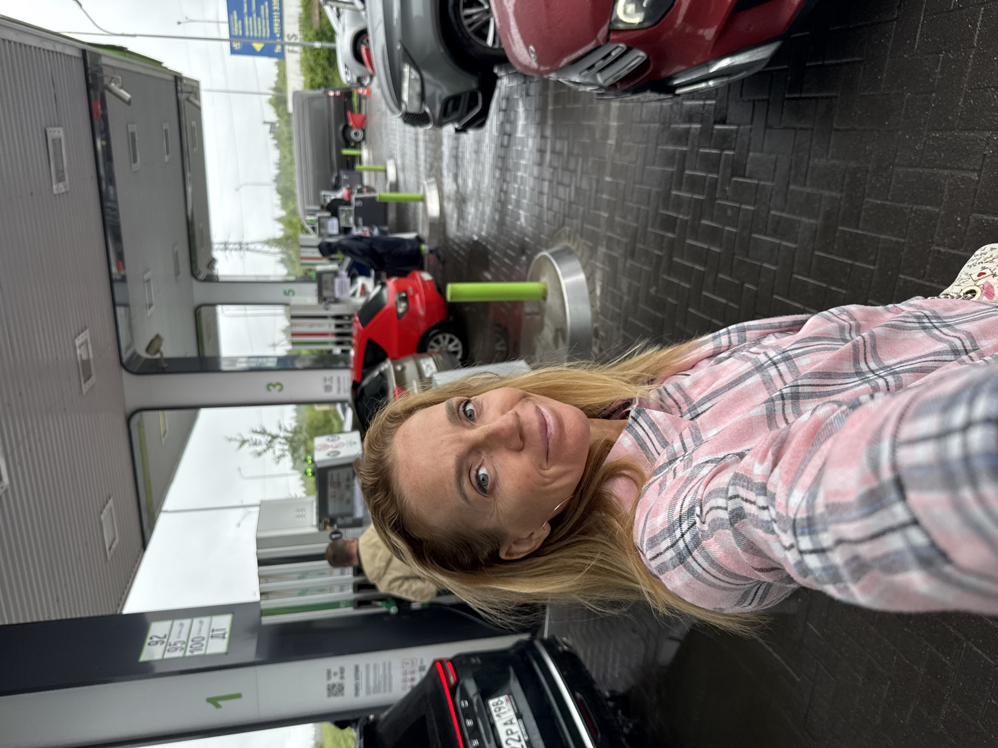 A blonde woman wearing a pink and white plaid shirt takes a selfie in front of a gas station in Medovoye, Russia. She is standing in front of a black SUV, and other cars, gas pumps, and a gas station canopy are visible in the background. The ground is wet and reflects the surrounding lights. There are informational signs with Russian text at the gas station.
