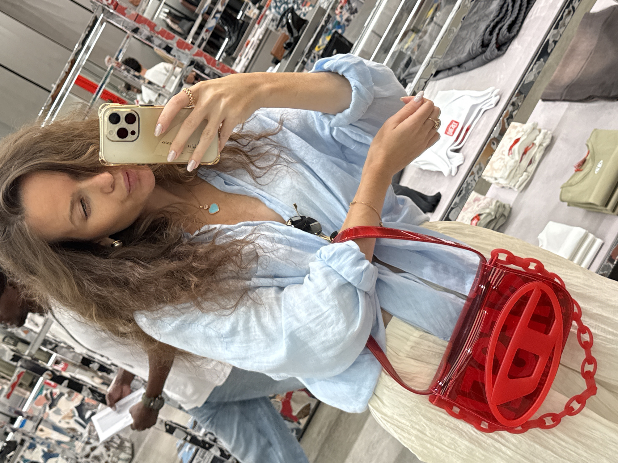 A woman with long, wavy brown hair takes a selfie in a retail store. She is wearing a light blue, oversized button-down shirt, a light-colored pleated skirt, and a red transparent handbag with a large, red Diesel logo on the front and a chain strap. A man in a white t-shirt and blue jeans is visible in the background, holding a piece of paper. The store appears to be a clothing store, with racks of clothes and shelves displaying merchandise. The environment is brightly lit, with the reflection of shelves and clothing visible in the mirrored surfaces of the store. The woman is holding a gold iPhone in her right hand, and wearing rings on her right ring finger and a bracelet on her left wrist. She has a light blue heart-shaped pendant necklace.