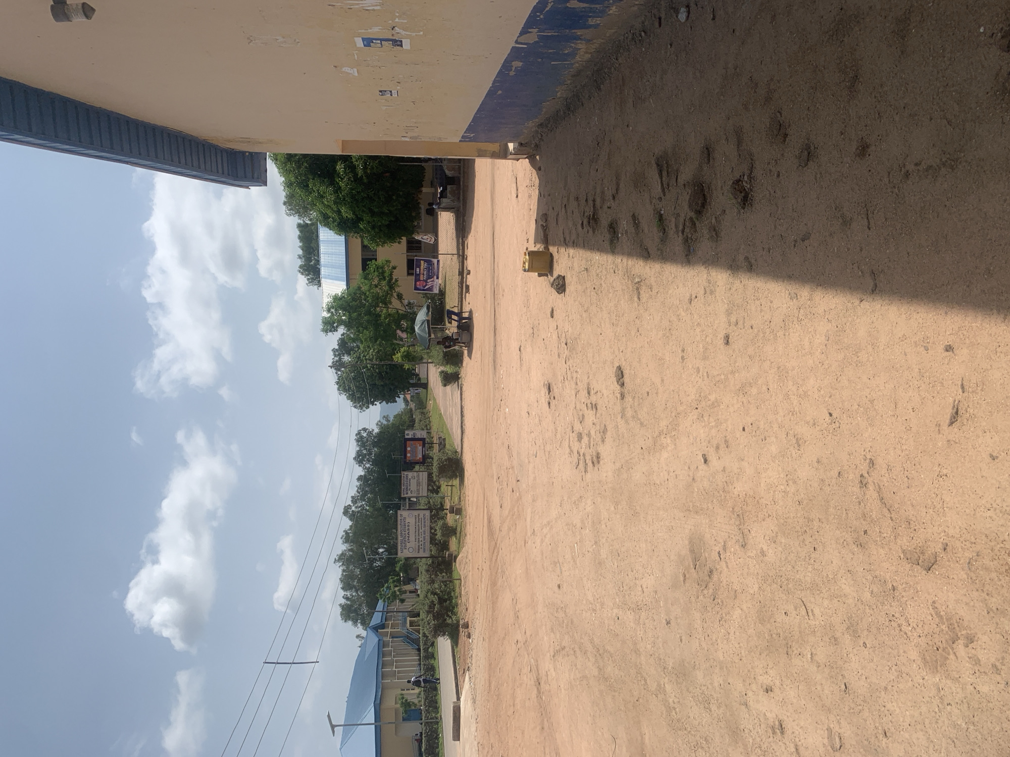 The image depicts an outdoor scene on a dirt road in Jalingo, Nigeria, likely on a university campus given the presence of institutional buildings and signage. In the foreground, the ground is predominantly dry, dusty earth with scattered stones and vegetation. To the left, a light-colored building with a blue roof and an overcast sky can be seen. In the mid-ground, there are several signs with text, including one that reads "NATIONAL CONVOCATION OF STATISTICS STUDENTS (NACOSS)". Two individuals are visible near a stall with a green umbrella. Further in the background, more buildings and trees are present, with a mountainous landscape in the distance. A person is walking on a paved path in front of the building on the left. On the right side of the image, a portion of a tan-colored building with a blue stripe near the bottom is visible, its wall appearing somewhat weathered. Overhead power lines traverse the scene. The overall impression is a bright, sunny day with scattered clouds.