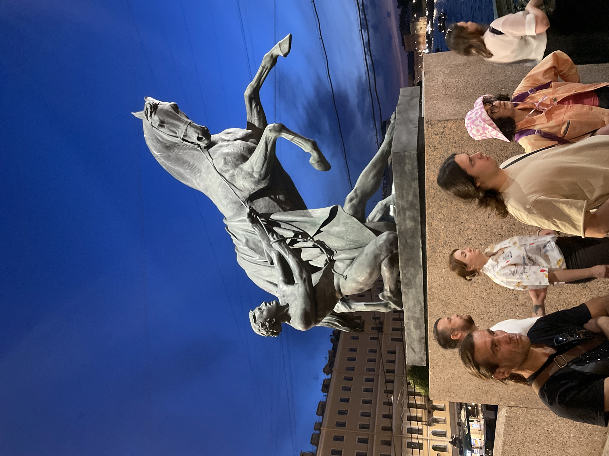 This evening scene depicts one of the "Horse Tamers" statues on the Anichkov Bridge in Saint Petersburg, Russia, with the Fontanka River visible in the background. The sky is a deep twilight blue, transitioning from lighter overhead to a darker, almost purple hue near the horizon, with faint cloud wisps.
The central subject is a large bronze equestrian statue, showcasing a powerful, muscular male figure on one knee, straining to control a rearing horse with ropes. The horse's front legs are elevated, and its head is turned slightly to the right, conveying immense struggle and dynamic motion. The statue rests on a solid, dark stone plinth.
In the mid-ground, a light-colored, multi-story building with numerous windows lines the left side, behind the statue. To the right, beyond the statue, a body of water with distant city lights and reflections indicates the Fontanka River. Overhead power lines are visible against the sky.
In the foreground, several individuals stand on what appears to be the bridge's stone structure. From left to right:
* A man with long brown hair tied back, wearing a black t-shirt and a brown shoulder bag, stands with crossed arms, looking intently to his right.
* Behind him, a bearded man in a white t-shirt stands in profile, facing right.
* Next, a woman with brown hair pulled back, wearing a light-colored, patterned short-sleeved shirt and dark pants, faces right, seemingly observing the river.
* Adjacent to her, a young man with long brown hair, wearing a light beige t-shirt, also faces right.
* To his right, a woman with glasses and dark hair wears a pink and white patterned bucket hat, an orange jacket, and a purple backpack, looking towards the statue.
* Farthest right, a woman with light brown hair, dressed in a white blouse and dark skirt, stands in profile, looking towards the river.
The overall lighting is dim, characteristic of the blue hour or late dusk, with the statue primarily silhouetted against the brighter sky, while the foreground figures are softly illuminated by ambient city lights.