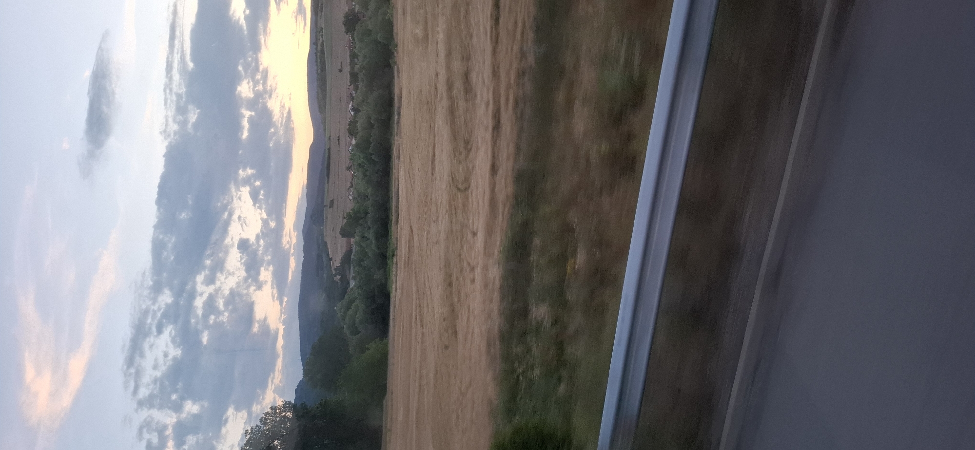 An outdoor, eye-level shot captures a rural landscape at dusk, possibly in Blajievo, Bulgaria. The foreground features a grey metal guardrail that runs horizontally across the bottom third of the image, casting a subtle shadow on the dark asphalt road below. Beyond the guardrail, a field of dry, golden grass, likely wheat or barley, stretches into the middle ground. Subtle tire tracks are visible in the field, suggesting recent agricultural activity or vehicular passage. A line of dense, dark green trees forms a visual barrier between the field and the distant rolling hills. Nestled amongst the trees and on the hillsides are scattered houses with red roofs, indicating a small village. The sky above is a soft blend of pale blue and pink hues, with scattered clouds that are tinged with the warm light of the setting sun. The overall impression is one of serene, rural tranquility at the close of day.