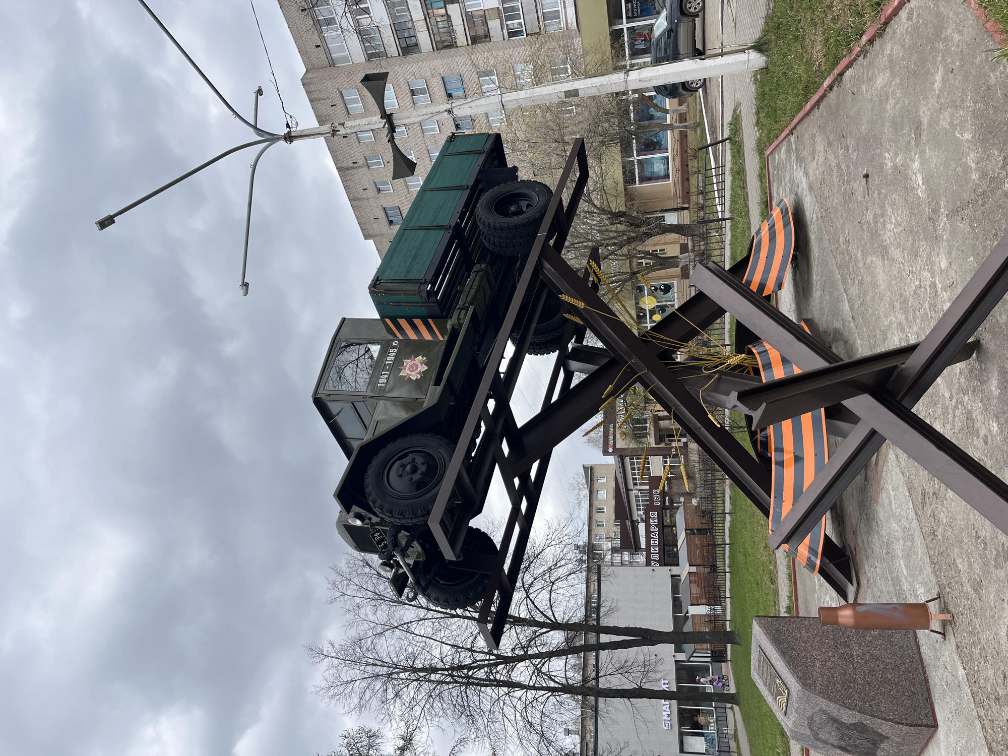 The image shows a memorial in the city of Chaikovsky, Russia. A green military truck is mounted on a metal structure resembling anti-tank obstacles. The structure is decorated with black and orange striped ribbons. A stone pedestal with a plaque and a replica artillery shell casing are visible at the base of the memorial. Buildings are in the background. The sky is overcast.