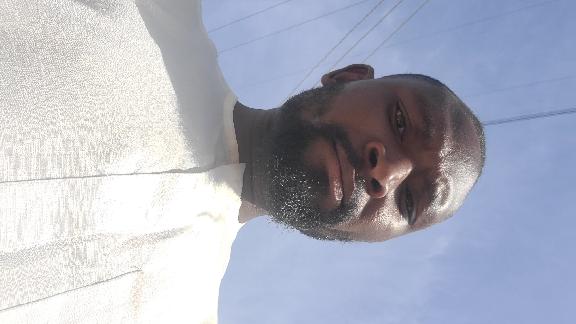 A medium close-up shot looking up at a Black man with a full beard wearing a light-colored, textured shirt. The background is a clear, light blue sky. Overhead power lines are visible in the background. The location is unspecified within Barakin Akawo, Nigeria.