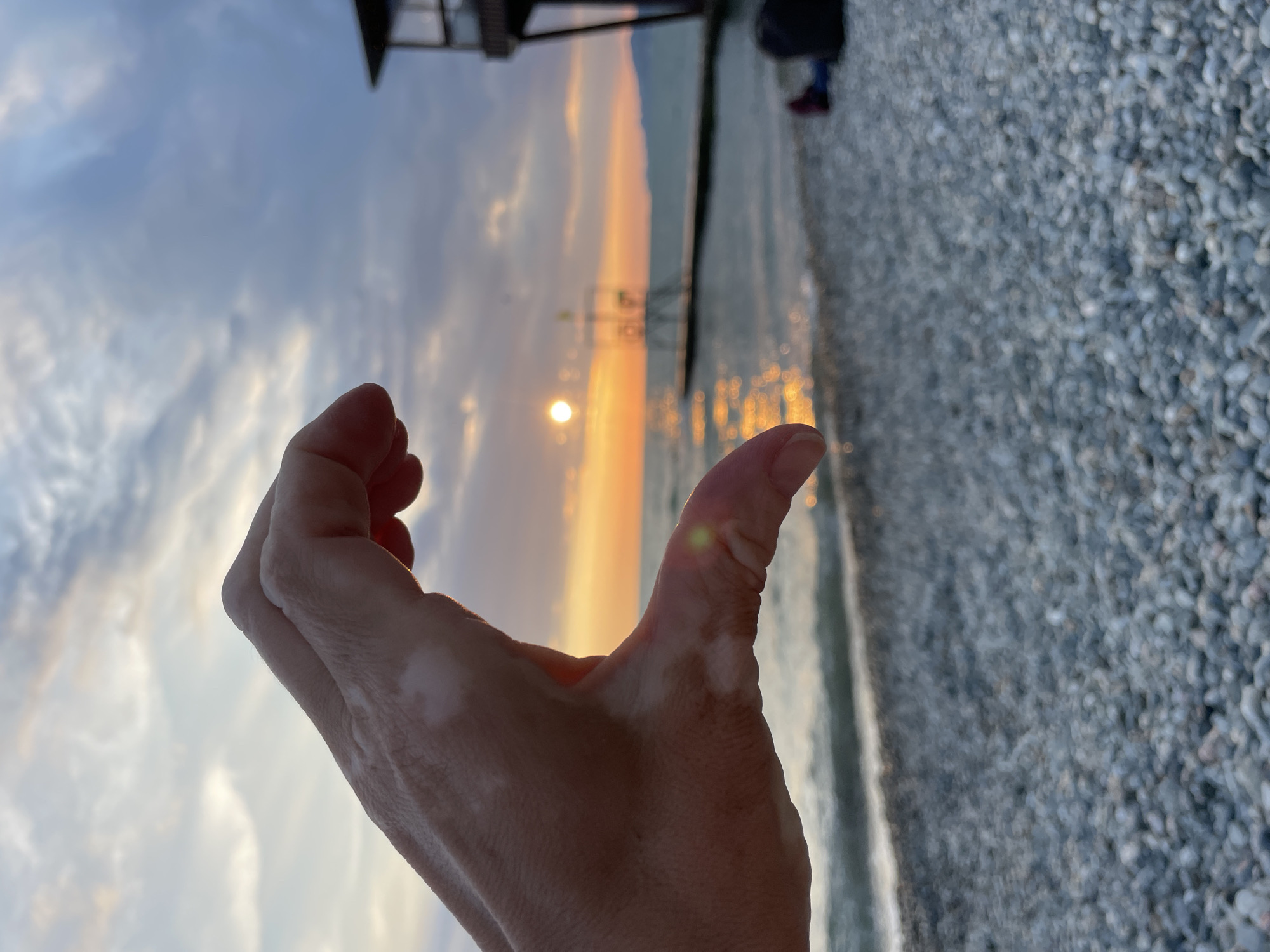 The image is a close-up shot of a person's hand in the foreground, forming a crescent shape. The hand is positioned against a blurred background depicting a beach scene at sunset. The sun is low on the horizon, casting a warm orange glow over the sky and reflecting on the water. A pier extends into the sea, with a small structure at its end. The beach is covered in pebbles. In the far right of the background, a person is sitting on the beach, with their legs extended. The sky is partly cloudy. The location is specified as Khostinskiy, Russia.