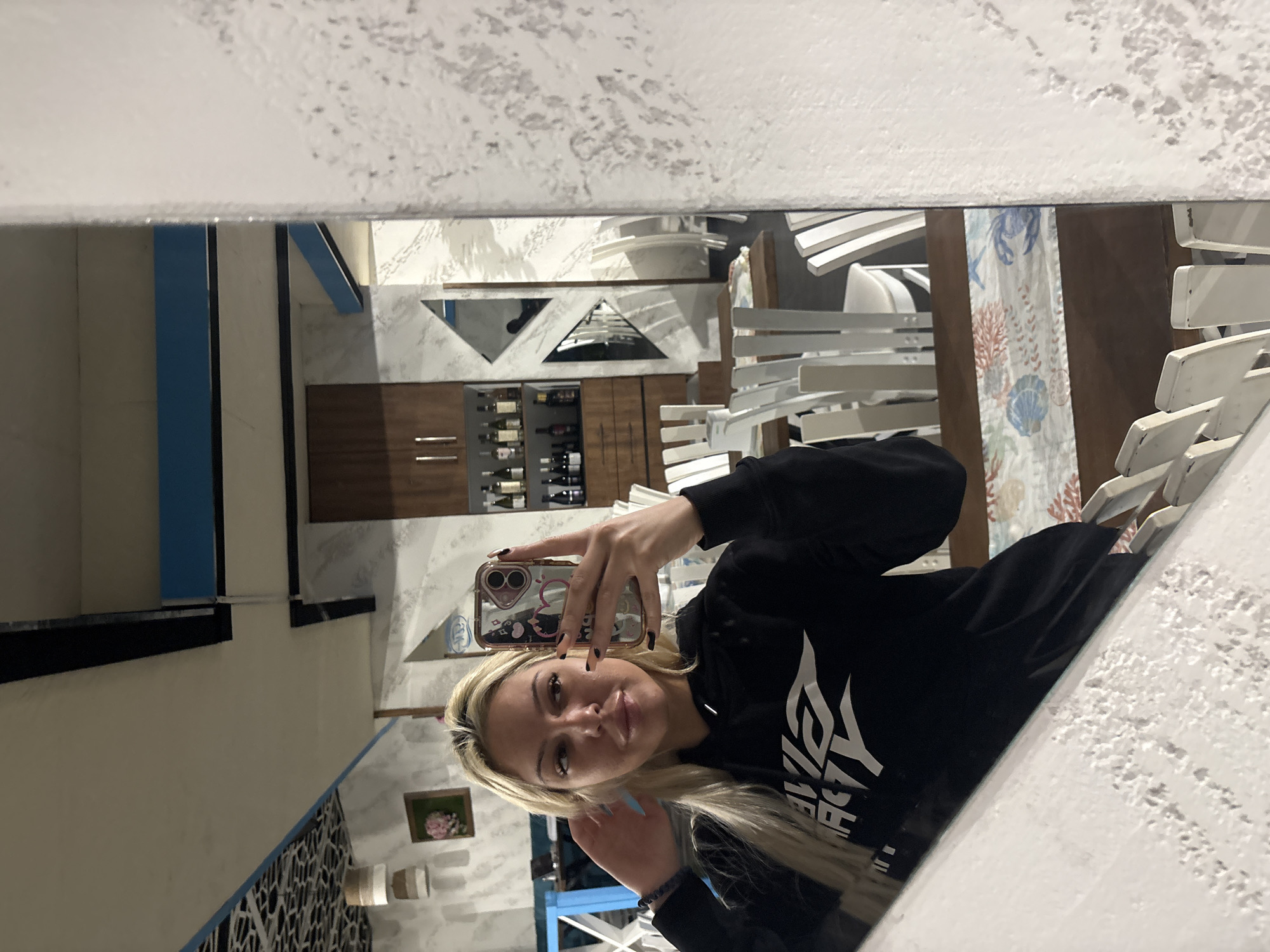A young woman with long blonde hair is depicted taking a mirror selfie, appearing rotated ninety degrees counter-clockwise from an upright position. She looks directly at the camera with a neutral expression, holding a smartphone with a reflective case in her right hand. Her left hand is near her hair. She wears a black hoodie with white text on the front, which partially reads "ALPHA ENERGY," and has black nail polish on her fingers. A dark beaded bracelet is visible on her left wrist.
The mirrored reflection reveals an indoor setting, likely a restaurant or dining area in Plovdiv, Bulgaria. The background features several white, slatted chairs and tables. Decorative elements include wall-mounted triangular mirrors, a small framed picture containing pink flowers, and a textured white wall. A dark wooden cabinet or shelving unit, stocked with various bottles, is also visible. On one of the tables, there is a long fabric runner adorned with a marine-themed pattern featuring a crab, coral, and a seashell. Blue and white architectural elements or columns can be seen in the reflection. The ceiling appears white and textured. The lighting suggests an indoor environment, possibly artificial or evening light.