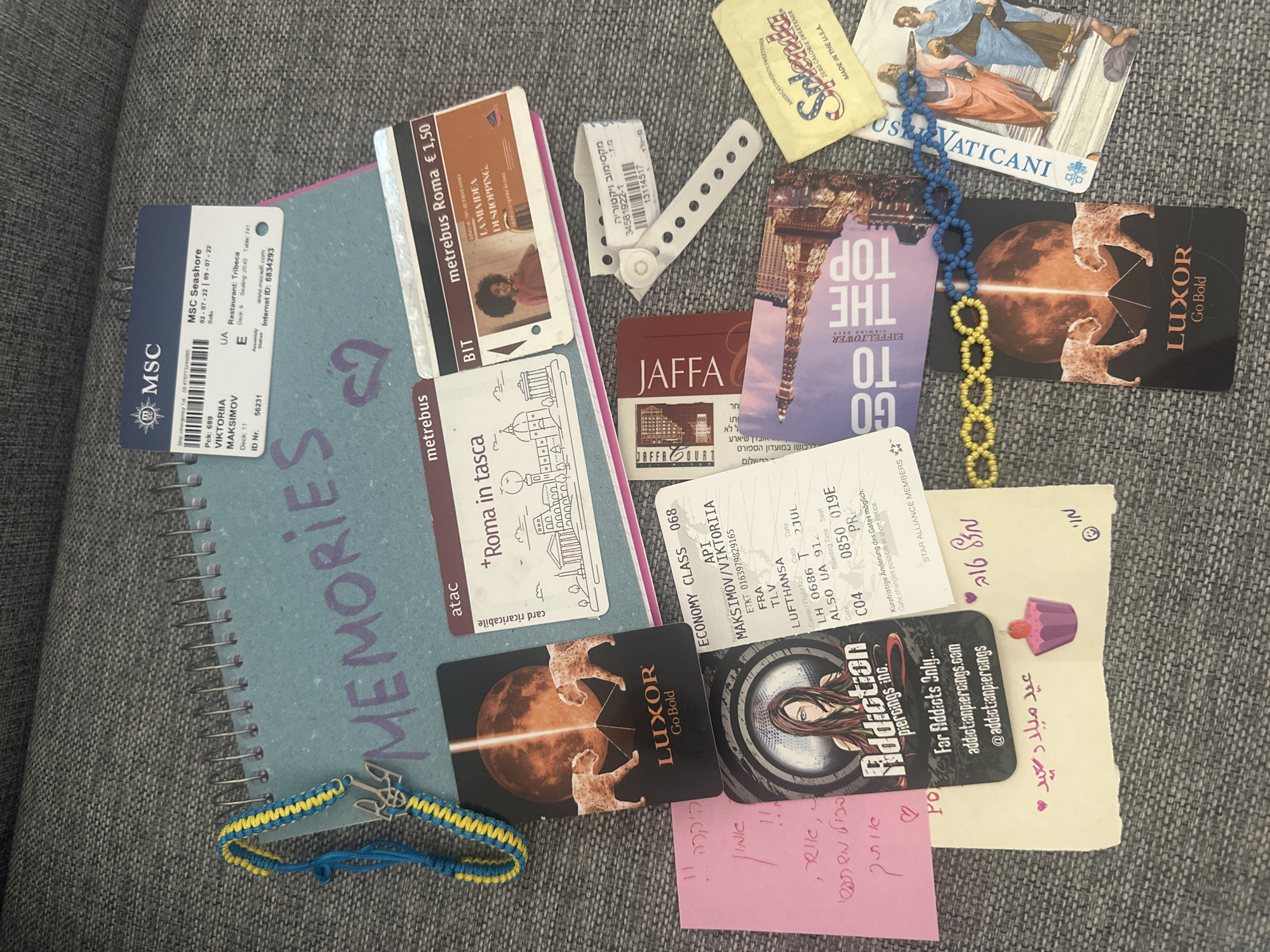 The image shows a collection of travel memorabilia arranged on a gray fabric surface, possibly a couch. A spiral-bound notebook with the handwritten word "MEMORIES" serves as a backdrop. Several tickets and cards are scattered around the notebook, including metro tickets for Rome, Italy ("metrebus Roma € 1,50"), a cruise ticket for MSC Seashore, and a Lufthansa boarding pass for a flight from Frankfurt (FRA) to Tel Aviv (TLV) for a passenger named Maksimov/Viktoriia.
There are cards from Luxor Go Bold, tourist information cards for Jaffa, Israel, and a card for the Eiffel Tower viewing deck. A small packet of Splenda sweetener and a card with a religious image are also present. There is also a blue and yellow Ukrainian friendship bracelet. There are also handwritten notes, including a birthday greeting in Arabic ("عيد ميلاد سعيد"). A business card for Addiction Piercings Inc. is also visible.
There's a wristband with Hebrew text and numbers (possibly a hospital or event wristband). There is no indication that the items are located in Petah Tikva, Israel.