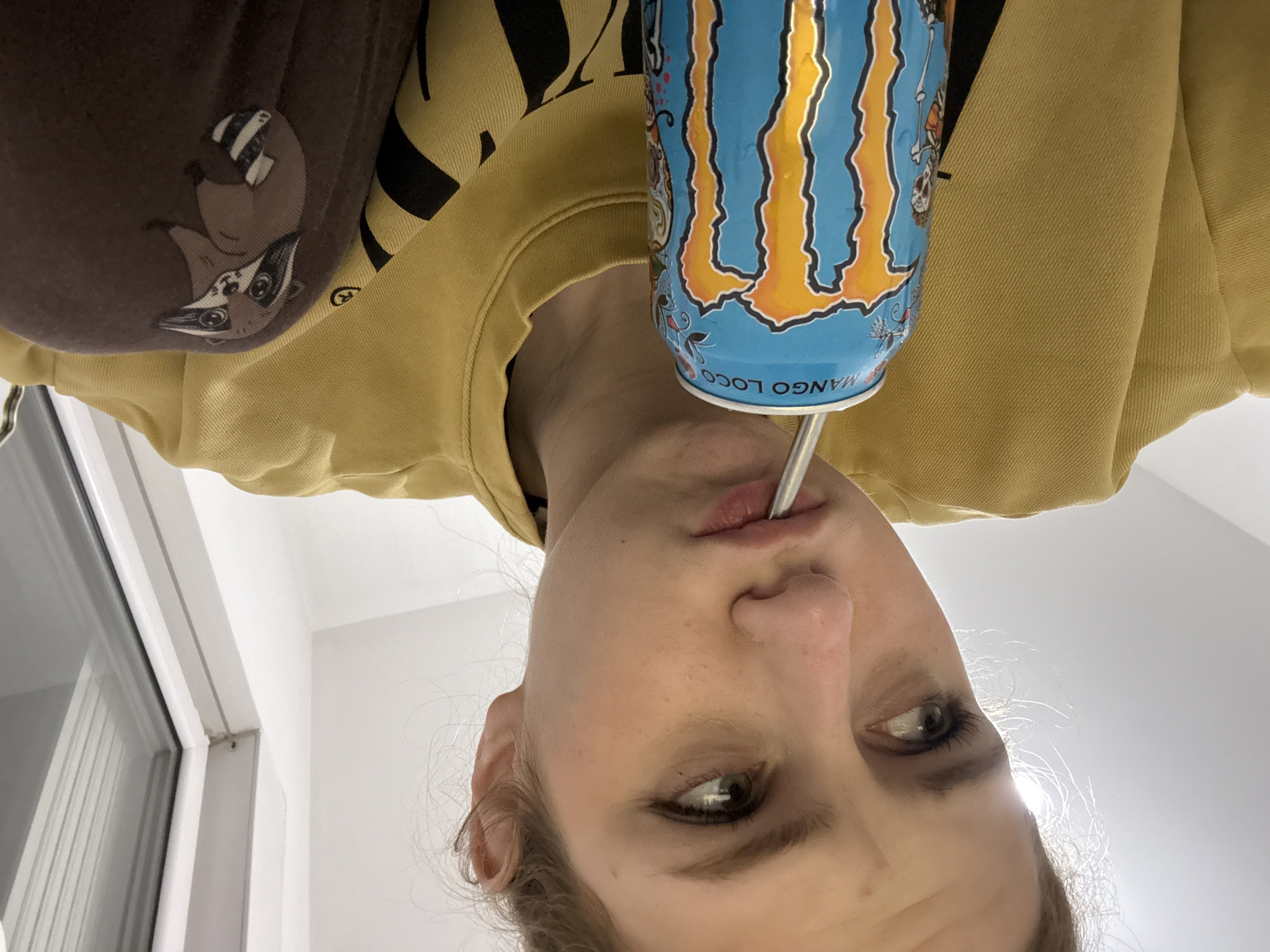 A person is drinking from a Mango Loco Monster energy drink using a straw. The person is wearing a mustard-colored sweatshirt with black lettering and a brown article of clothing with a cartoon raccoon on the shoulder. The photo appears to have been taken indoors, based on the lighting and window, but the location cannot be determined from the image. It cannot be ascertained if the location is Kragujevac, Serbia.