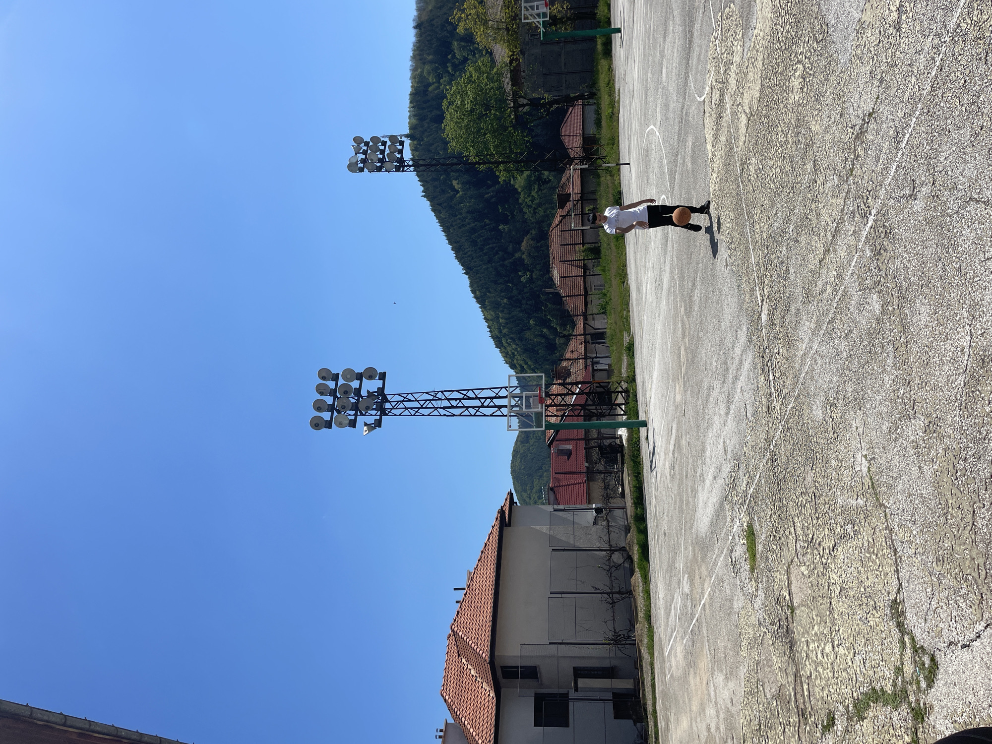 A basketball court is located in Zlatograd, Bulgaria, on a clear, sunny day. A person in a white shirt and dark pants is dribbling a basketball. Tall light posts stand near the court. The court's surface is cracked and weathered. Buildings with red-tiled roofs and a wooded hillside are visible in the background.