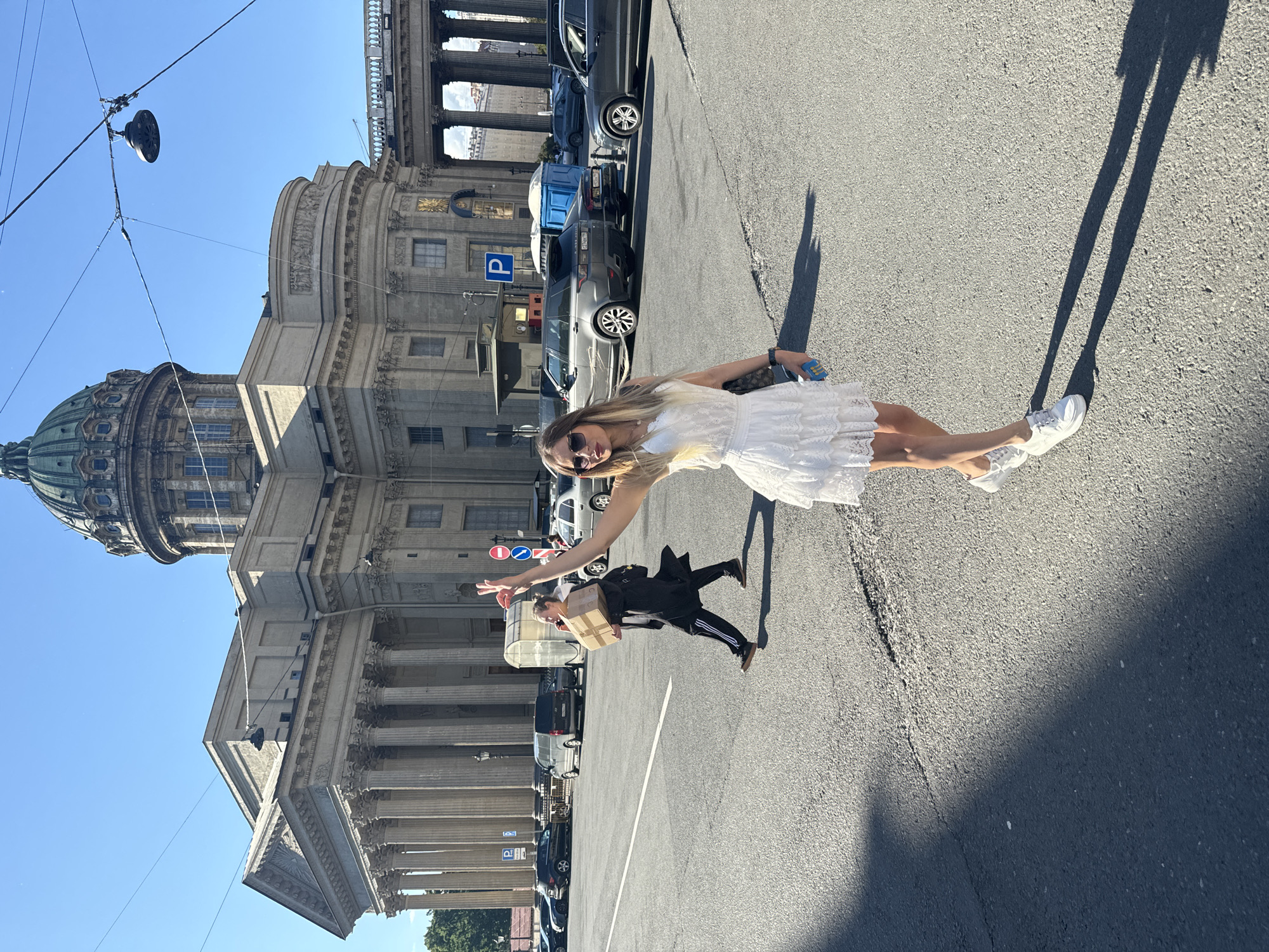 In Saint Petersburg, Russia, a woman in a white dress and white sneakers poses for a photo, holding a blue object in her right hand and making a peace sign with her left. Behind her, a person in a black coat and black pants, also in motion, carries a cardboard box. The scene is set on a sunlit street with a large, classical-style building with columns and a dome in the background. Several cars are parked and driving on the street, and traffic lights and signs are visible. The overall environment suggests an urban setting with notable architecture.