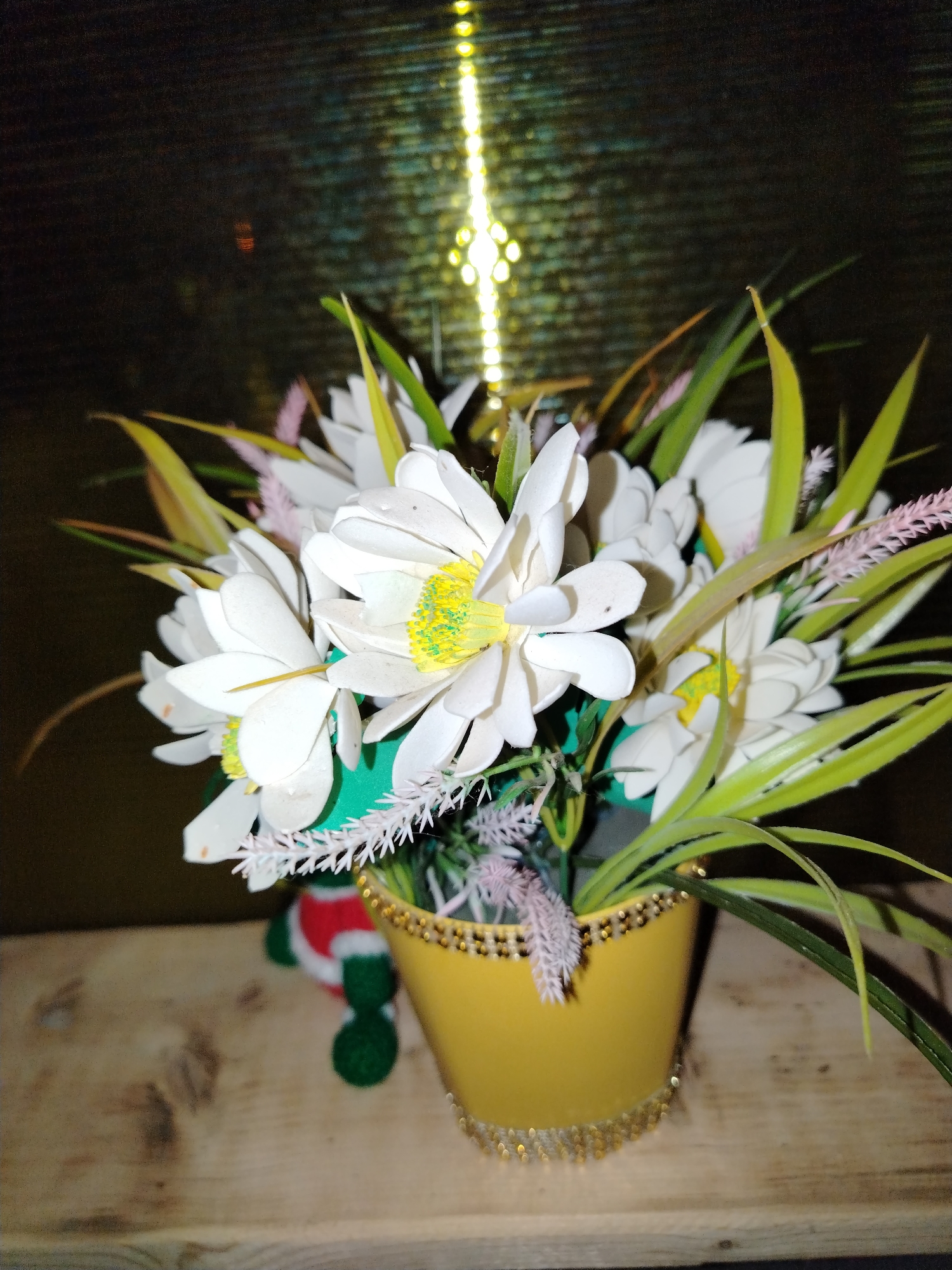 The scene depicts an artificial floral arrangement positioned on a wooden surface. The arrangement consists of a yellow pot, adorned with two horizontal bands of golden, spherical embellishments at its rim and near its base. Contained within are multiple artificial white flowers, characterized by bright yellow-green centers, intermingled with elongated green leaves, light pink-purple feathery stalks, and white spiky decorative elements. A green cylindrical object is partially visible within the flower cluster. To the left and slightly behind the pot, a blurred, red and green fabric figure, resembling a decorative gnome, is partially visible. The wooden surface beneath the pot displays visible grain. The background is dark and out of focus, featuring a prominent vertical strip of diffused, yellow-orange light sources on the right side. The location is Гожан, Russia.