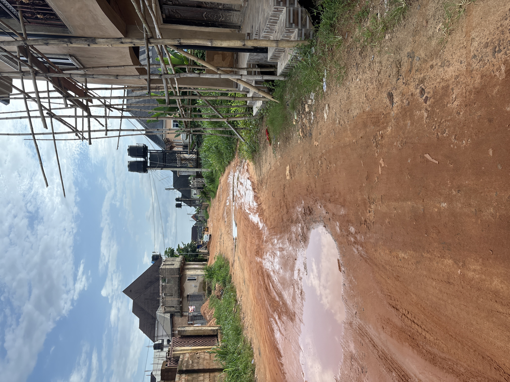 The image depicts a red dirt road in Obosi, Nigeria, heavily impacted by recent rainfall, evidenced by large puddles and mud. The road is lined with buildings in various stages of construction, characterized by scaffolding made of bamboo and unfinished facades. On the right side of the road, a building is under construction, with scaffolding providing access to its upper levels and a tiled staircase leading to the entrance. On the left side, more buildings are visible, some with completed roofs and others with exposed concrete structures. Residential water tanks are perched on top of some buildings. The sky is partially cloudy, with a mix of blue sky and white clouds. The overall scene suggests an active construction area in a developing urban environment.
