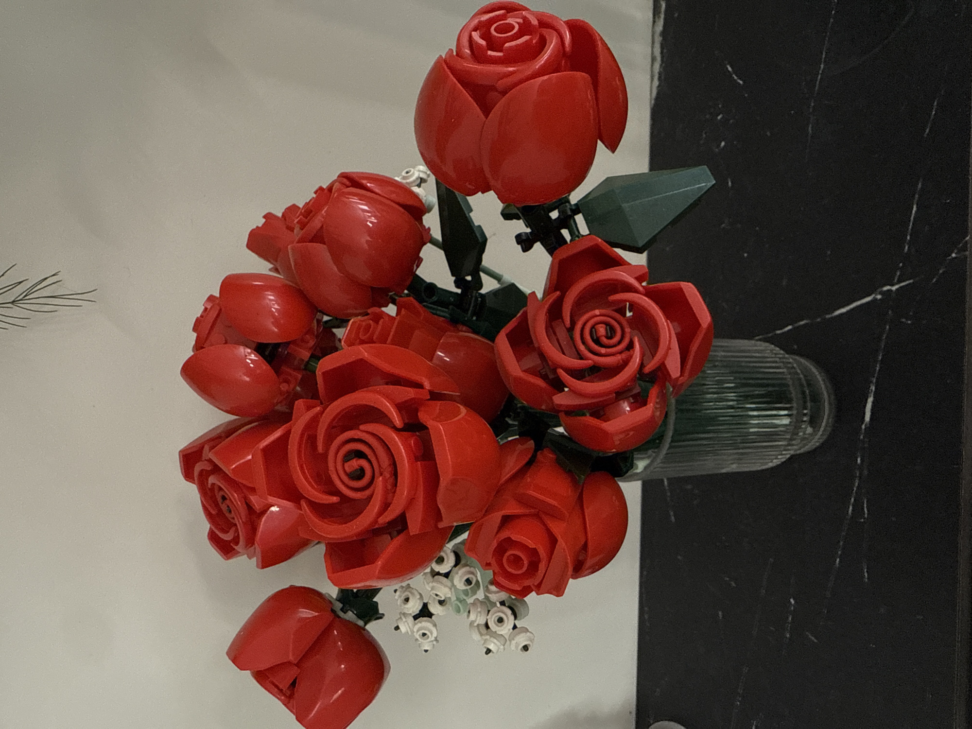 A bouquet of red roses, constructed from Lego bricks, is displayed in a ribbed glass vase. The vase sits on a black marble countertop against a plain white wall. The roses are a mix of open blooms and buds, with green Lego leaves and stems supporting them. To the left of the roses, a small cluster of white Lego flowers with black centers is visible. A delicate line drawing of a pine branch hangs on the wall above the bouquet. The scene is lit from the front, casting subtle shadows. The location is in the city of Khokhryaki, Russia.