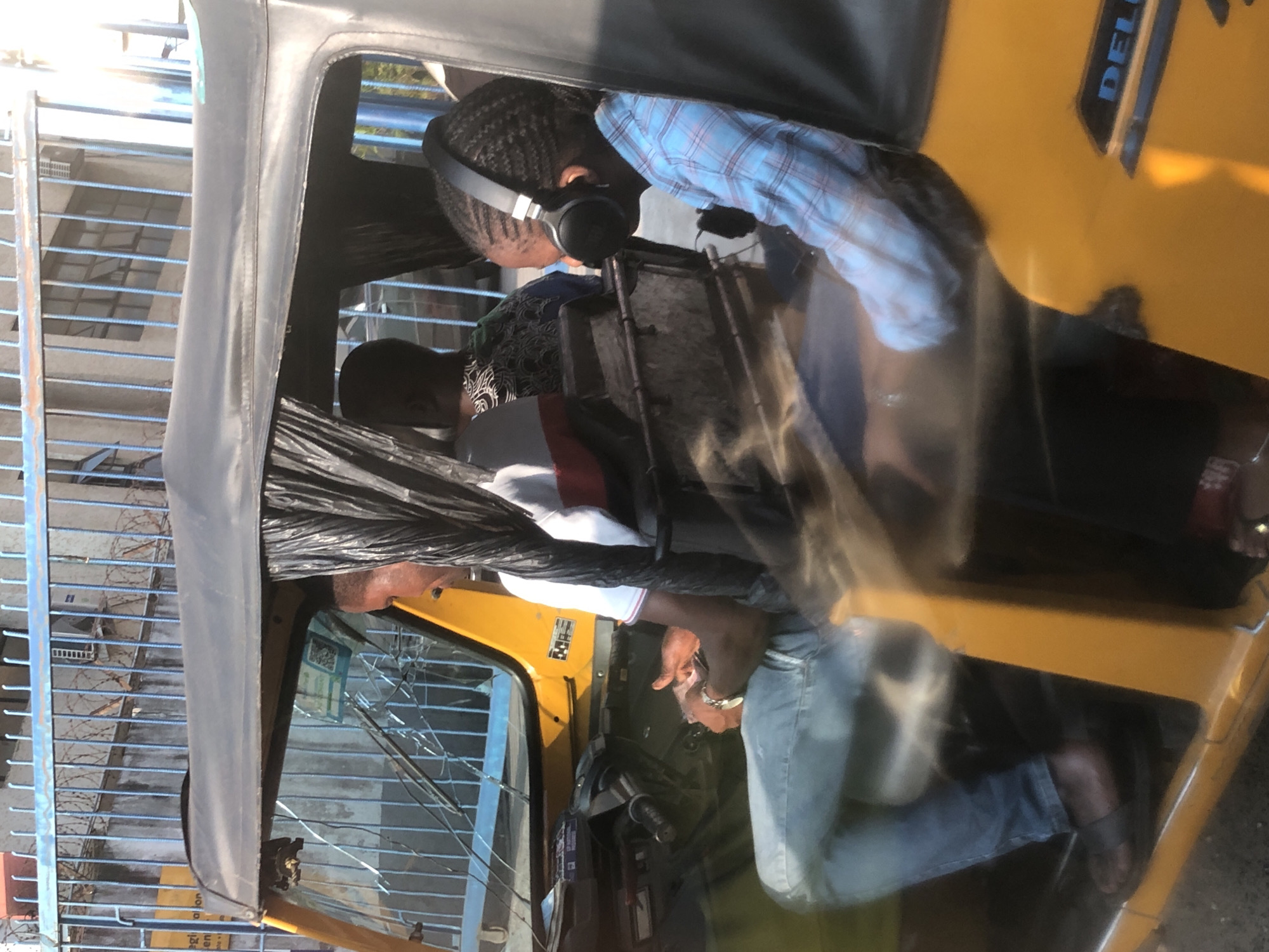 A front view of a yellow auto-rickshaw (Keke Napep) in Ikeja, Nigeria, with three occupants is captured. The driver, a man wearing a white t-shirt with red and blue stripes, blue jeans, and dark sandals, is seated in the front left. His hands are visible on the steering wheel. Behind him, a person wearing a patterned black and white shirt is seated. To the right of the driver, a young person wearing a blue and white plaid shirt is seated, listening to music with black headphones. The auto-rickshaw has a black fabric roof and a clear windshield. In the background, a blue metal fence with barbed wire and a building with windows are visible. The scene appears to be on a street in Ikeja, Nigeria, indicated by the presence of the auto-rickshaw, a common mode of transport in the city.