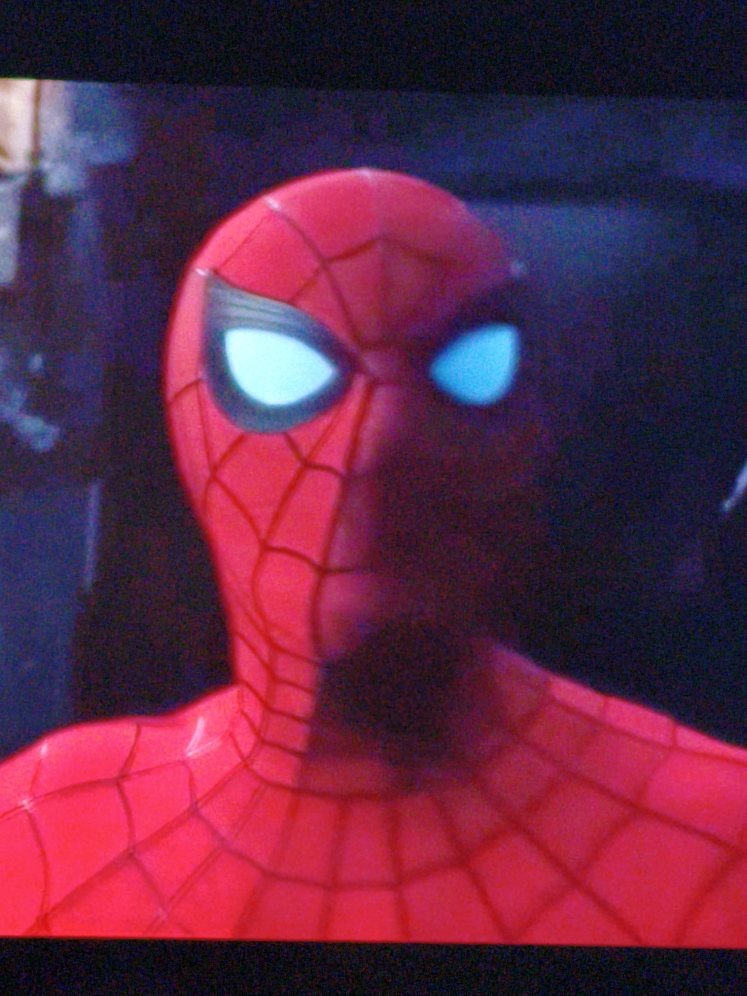 This is a close-up, low-angle, pixelated, and grainy digital photograph of a Spider-Man costume. The costume is predominantly red with a black web pattern. The iconic eye lenses of the mask are bright cyan and illuminated. The background is dark and out of focus, with some blurry blue and purple hues, suggesting an indoor or nighttime setting. There is no discernible indication of the city of Vladivostok, Russia, or any other specific location. No people, actions, or interactions are visible, only the costume itself.