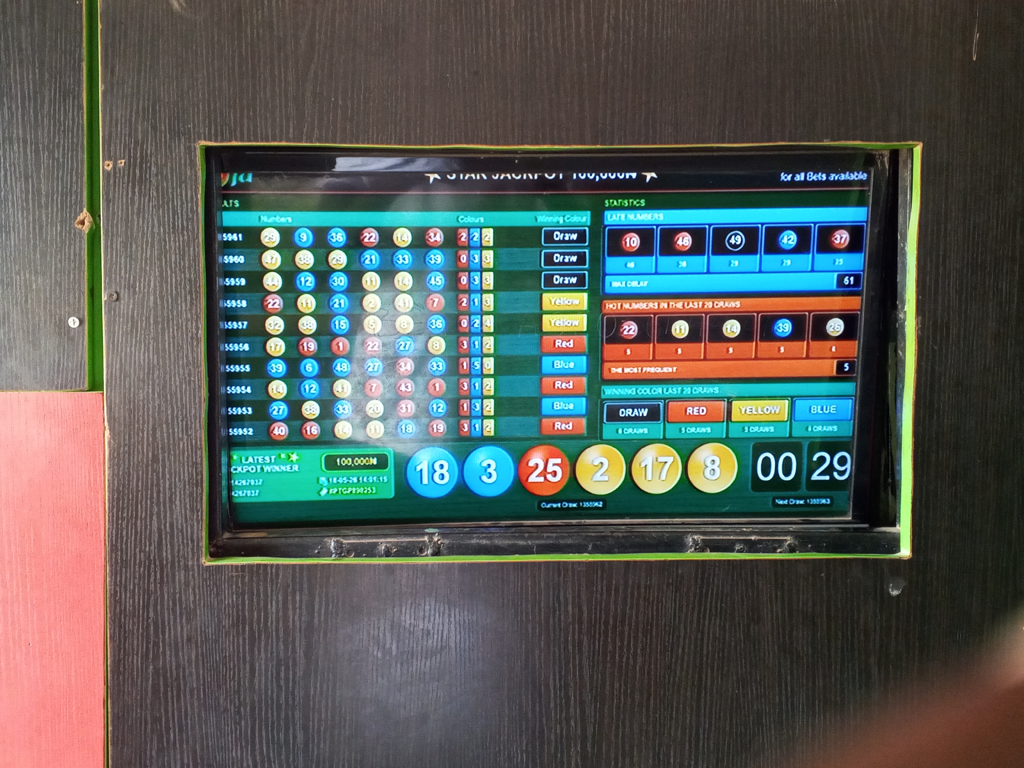A digital display is mounted on a dark brown wooden paneled wall, which is framed in black trim. The screen displays betting statistics for a lottery or similar game. The top of the screen reads "STAR JACKPOT 100,000N" with "for all Bets available" underneath. The screen displays various statistics including "LATE NUMBERS", "HOT NUMBERS IN THE LAST 20 DRAWS" and "WINNING COLOR LAST 20 DRAWS". The "LATEST CKPOT WINNER" is 100,000N, with the number 14267837 and 1257837 listed below. A timer at the bottom of the screen displays "00 29". The screen's text suggests this is a betting establishment, possibly in the city of Gwong, Nigeria, as indicated by the local currency (N, Naira). The dark wooden panels suggest it's an indoor environment. There are no people visible.