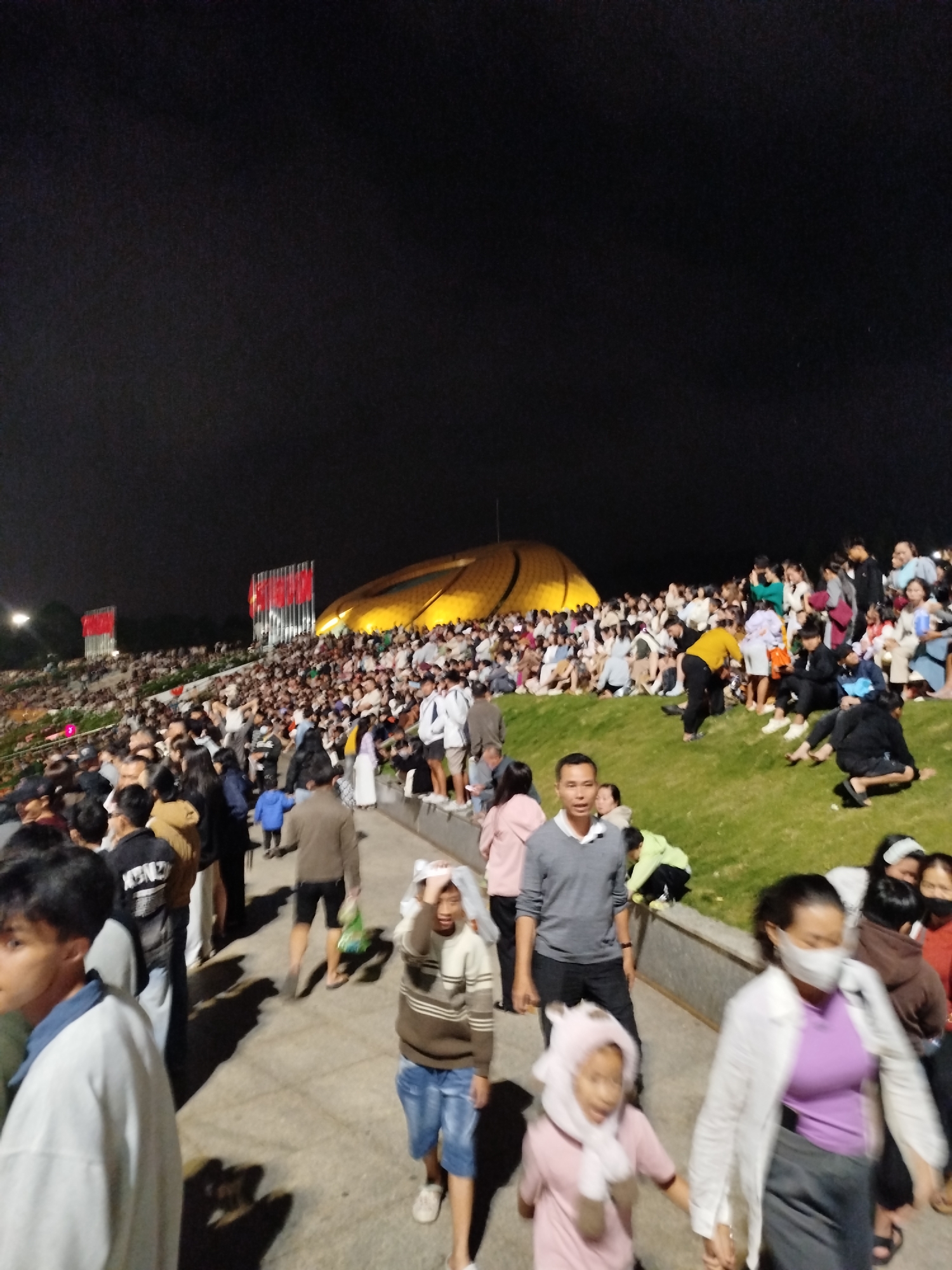 The image depicts a nighttime scene in Da Lat, Vietnam, likely during a public event or celebration. In the background, a large, illuminated, dome-shaped structure with a golden hue dominates the skyline, with two prominent red flags displayed on vertical structures in front of it. The foreground is filled with a large crowd of people of various ages, many of whom are standing or seated on grassy slopes. The people are dressed in casual attire, and some are holding what appear to be plastic bags. The ground appears to be paved in some areas and grassy in others. The lighting suggests an outdoor event taking place after dark, possibly a festival or fireworks display given the large gathering. The overall atmosphere seems to be one of a crowded public gathering under artificial light.