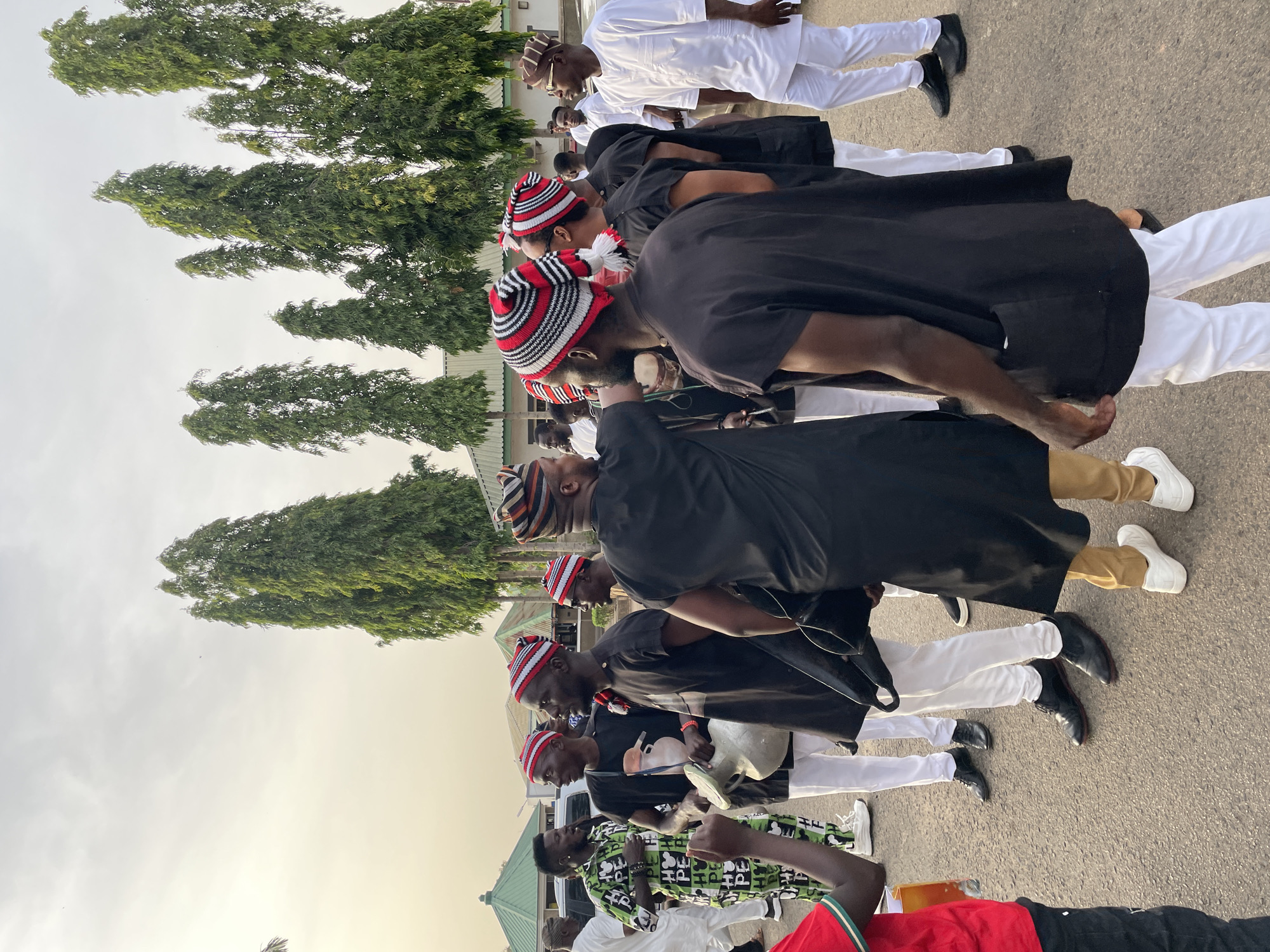 A medium outdoor shot depicts a group of men participating in a procession on what appears to be a paved street. The location is the city of Jos, Nigeria.
The men are arranged in a line, most wearing a similar attire consisting of white pants, black shoes, and black tops. A unique feature of their attire is a red, white, and black striped cap. Some men wear white attire with black shoes. The men are lined up next to tall trees.
The background includes trees and a glimpse of buildings. The sky is overcast, suggesting it might be dawn or dusk. There is one man wearing a green-and-white patterned shirt.