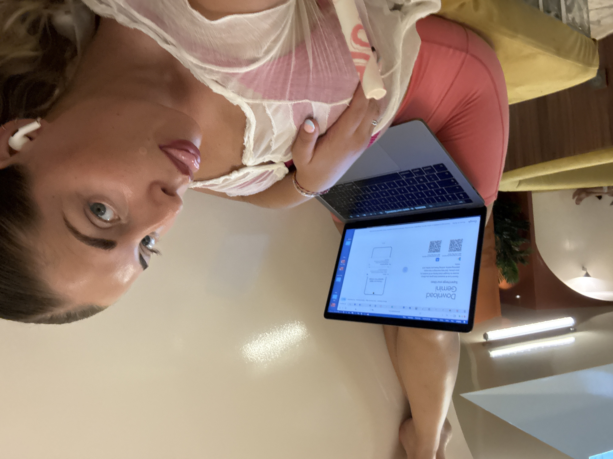 A woman is lying on her side, resting her head on her hand. She is wearing a white sheer top with pink fabric underneath, and pink shorts. She has blonde hair, blue eyes, and is wearing white earbuds. She is looking towards the camera with her tongue sticking out slightly. In front of her is an open laptop displaying a webpage with the words "Download Gemini" and two QR codes. The background shows a yellow sofa, a reflective surface, and some potted plants, suggesting an indoor setting. The overall lighting suggests it is daytime. There is no visible text other than on the laptop screen.