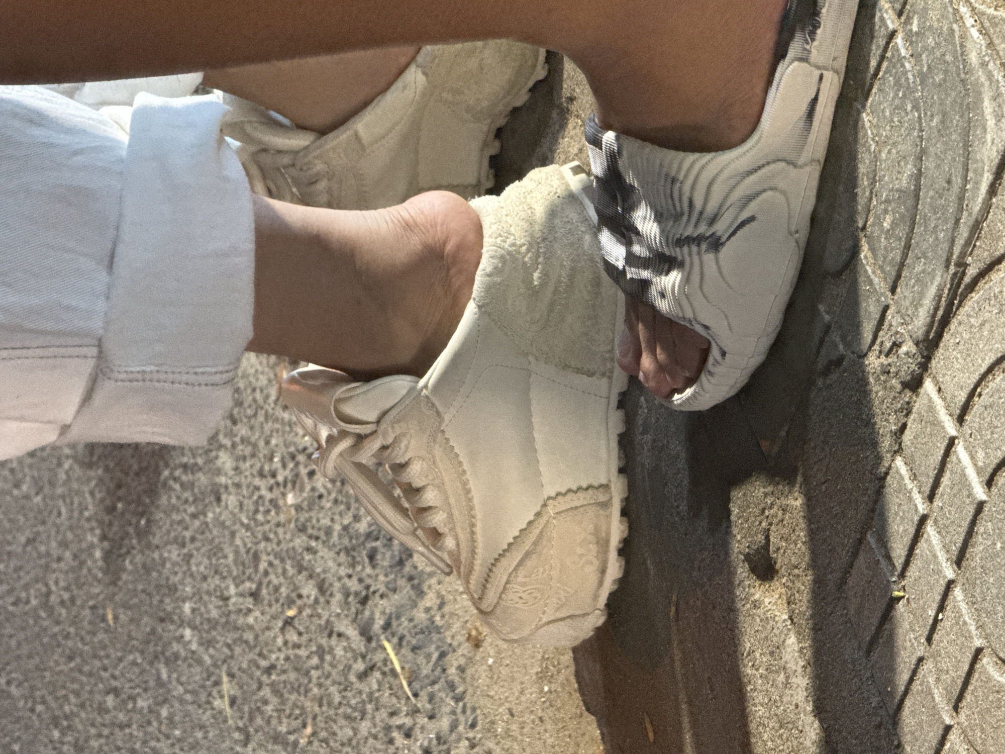 This close-up image, taken in Đà Nẵng, Vietnam, captures a person's lower legs and feet. One leg is clad in light-colored, possibly white or off-white, trousers with a neatly cuffed hem.
The person is wearing two different types of footwear. On the right, a foot is partially encased in a light-colored sandal or slide featuring a distinctive black and white, or grey and white, abstract, possibly marbled or striped pattern on its broad upper strap. The toes are visible through the sandal's open front.
On the left, the other foot is in a light-colored, likely beige or off-white, closed-toe shoe, resembling a sneaker or trainer. This shoe has visible laces, intricate stitching, and textured fabric panels on its sides. Around the ankle opening, there appears to be a fluffy or towel-like lining. The person's heel is slightly lifted from inside this shoe.
The ground surface beneath the feet is varied, comprising rough, speckled asphalt or concrete, alongside a section of textured paving stones arranged in a distinct, possibly leaf-like or rhomboid, pattern. Strong shadows are cast on the ground and footwear, indicating bright daylight conditions, suggesting either morning or late afternoon sun. No discernible text is visible in the image.