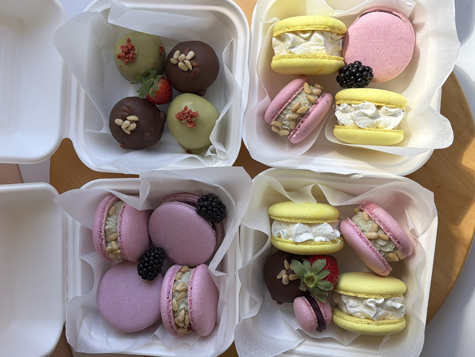 Four white, disposable takeout boxes filled with an assortment of macarons and spherical confections are arranged on a light-colored wooden surface.
**Box 1 (Top Left):** Contains five purple macarons. Two display a white, creamy filling topped with pine nuts, while the remaining three are solid-colored shells. Two black blackberries garnish the contents.
**Box 2 (Top Right):** Holds four spherical desserts and one red strawberry. Two spheres are dark brown and topped with pine nuts. Two spheres are light green and topped with small reddish-pink dried fruit pieces. The whole strawberry, with its green stem, is placed among them.
**Box 3 (Bottom Left):** Contains five macarons, one dark brown spherical dessert, and one red strawberry. Two macarons are yellow with a white, creamy filling. Two macarons are pink with a white, creamy filling and pine nuts. One small, solid dark pink macaron is partially visible. The dark brown sphere resembles those in Box 2. A whole red strawberry with its green stem is also present.
**Box 4 (Bottom Right):** Displays five macarons and one black blackberry. Two macarons are yellow with a white, creamy filling. One macaron is pink with a white, creamy filling and pine nuts. Two macarons are solid pink, one appearing as a full macaron and the other possibly a single shell. A single blackberry rests on a solid pink macaron.
All boxes are lined with white tissue paper. The image does not contain any visual information to determine a specific location within the city of Tel-Aviv, Israel.